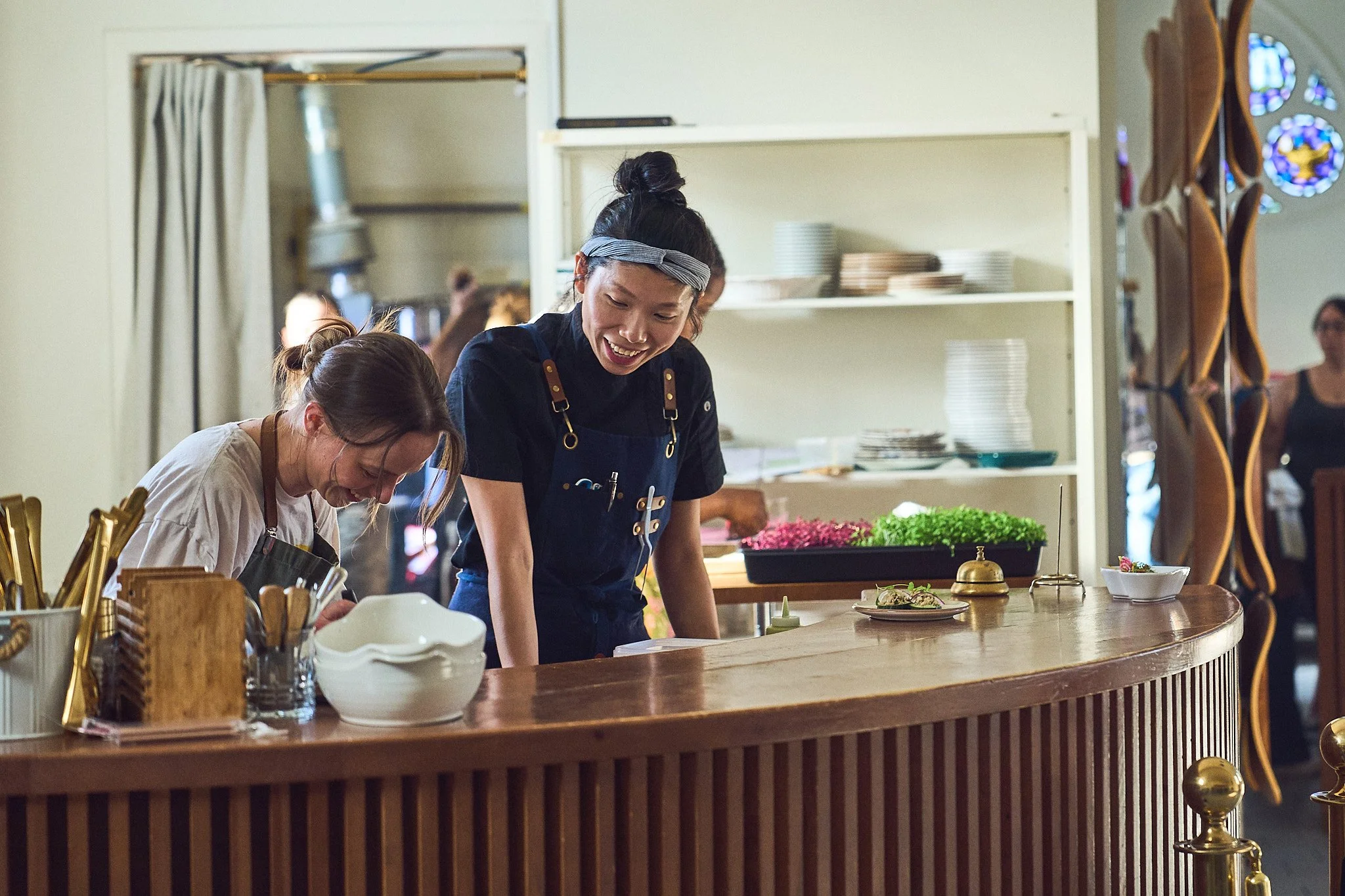 Nora Jo in a kitchen behind a wooden counter, surrounded by kitchen utensils and plates, with a person observing in the background at the Girl Dinner Event. Photographed by Gumption Studio, Ottawa event photographer.