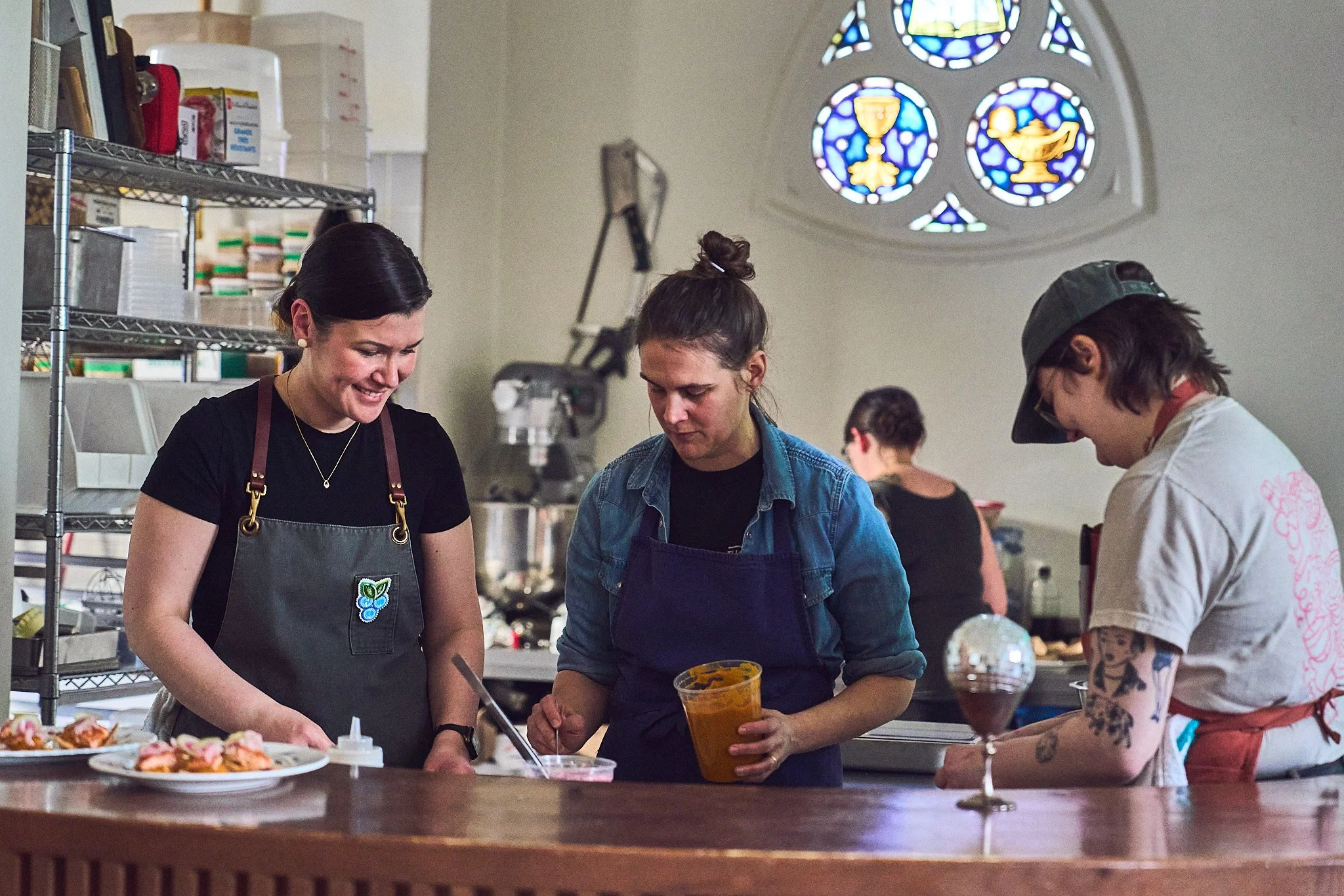 Three women working in a kitchen, preparing food and filling containers, with a stained glass window in the background. Photographed by Gumption Studio, Ottawa event photographer.