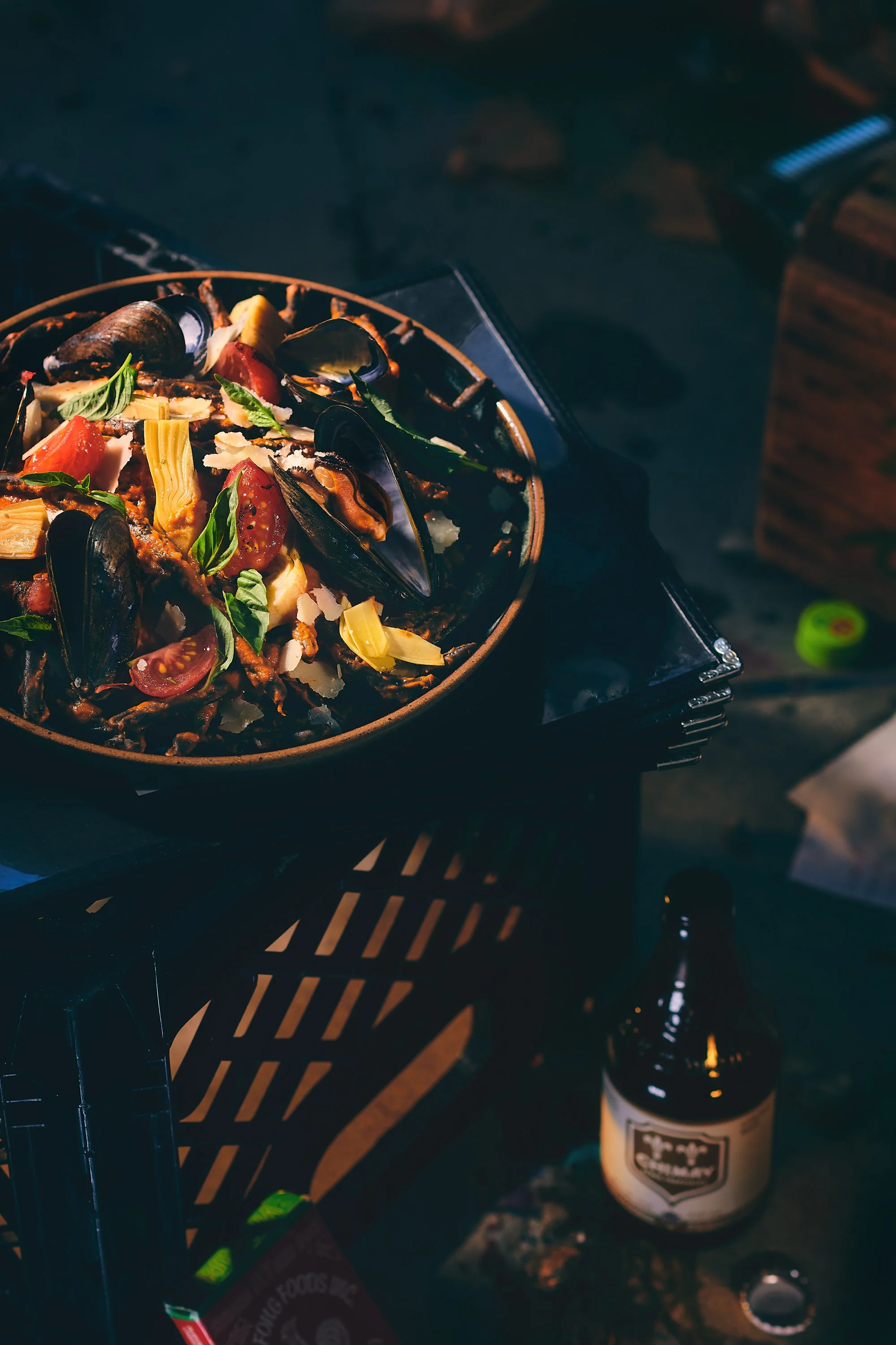 A pan of cooked pasta with cherry tomatoes, basil, cheese, and mussels on a grill table at an outdoor setting. A bottle with a label and a lighter are also visible near the pan. Photographed by Nick Ghattas, and Ottawa food stylist Gumption Studio.