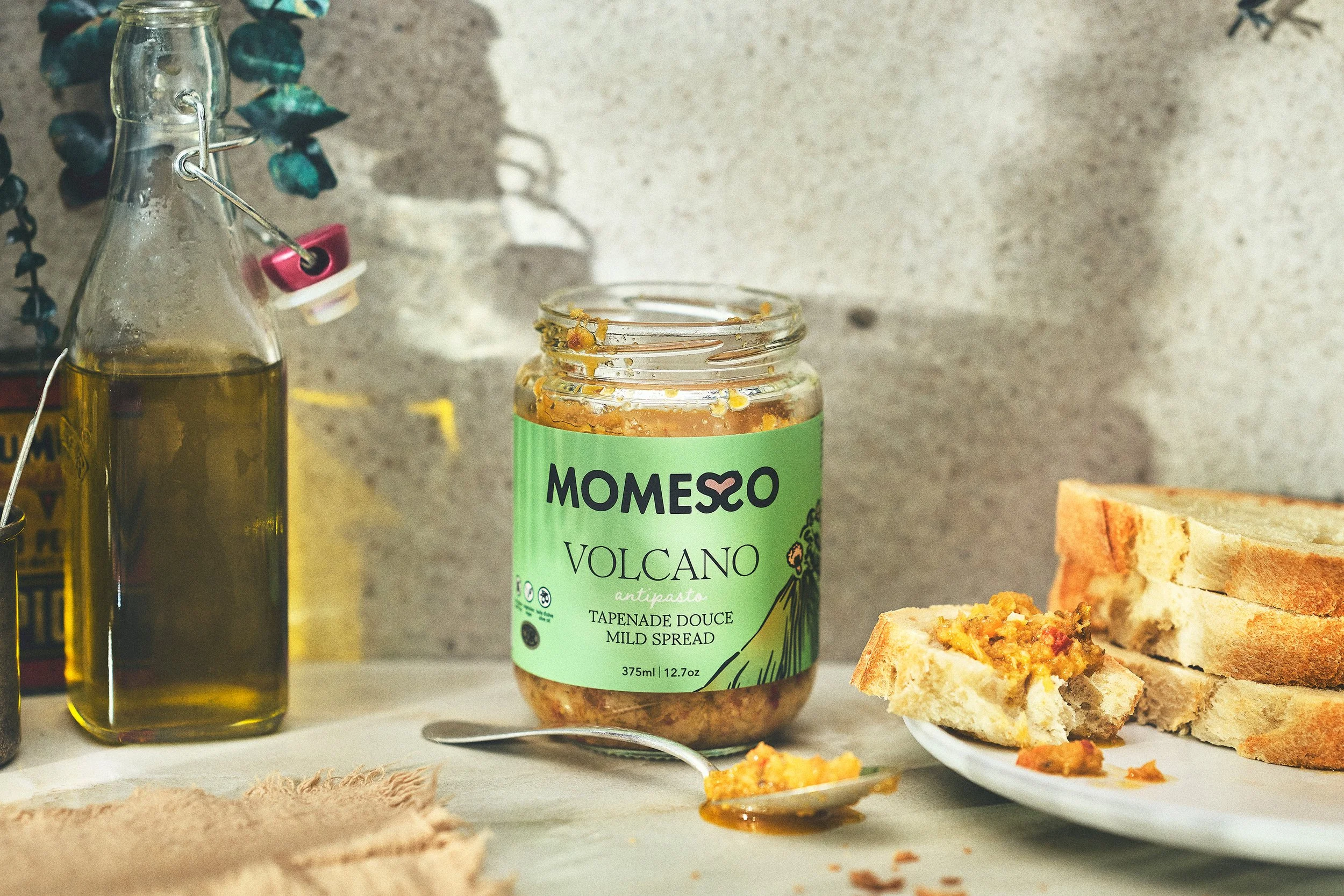 A jar of Momeso Volcano mild spread atop bread slices, with a bottle of olive oil, a spoon, and bread slices nearby, against a rustic background. Photographed by Nick Ghattas, and Ottawa food stylist Gumption Studio.