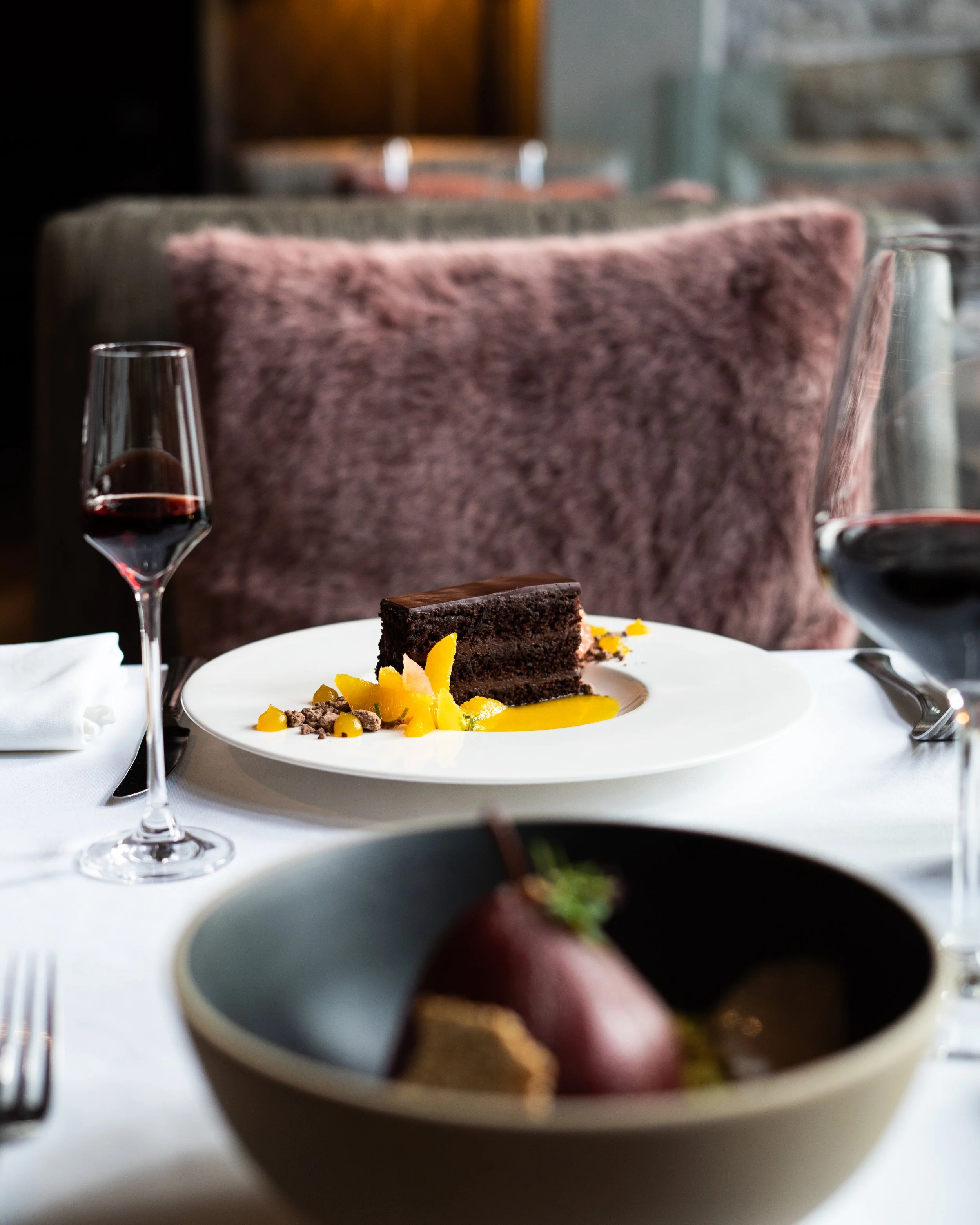 A dessert plate from Restaurant e18hteen with a piece of chocolate cake garnished with yellow fruit slices, served with a glass of red wine on a white tablecloth. Photographed by Gumption Studio, Ottawa food photographer. 