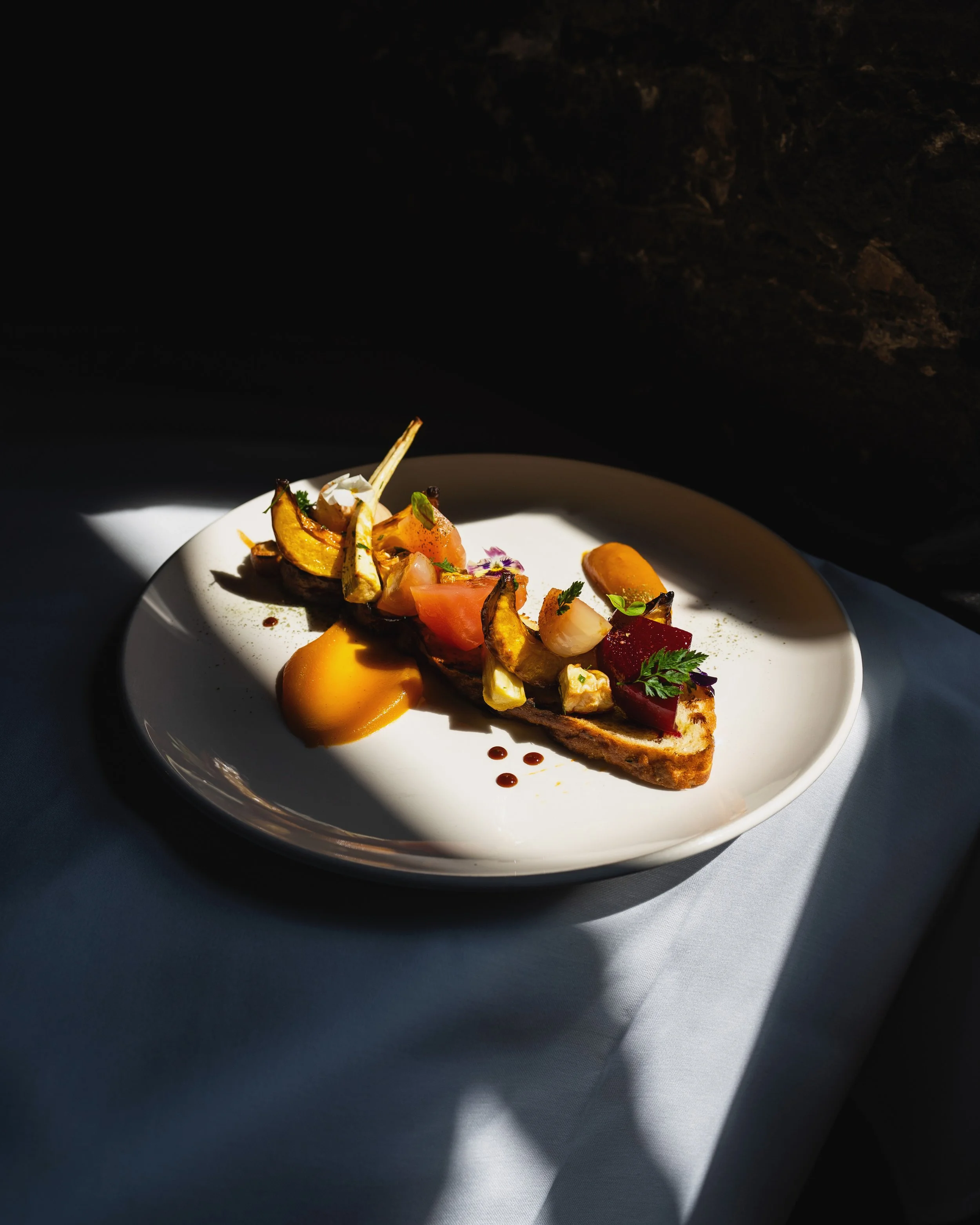 A dish presented on a white plate with vibrant garnishes, drizzled sauces, and carefully arranged ingredients, set on a white tablecloth with dramatic lighting casting shadows. Photographed by Gumption Studio, Ottawa food photographer.