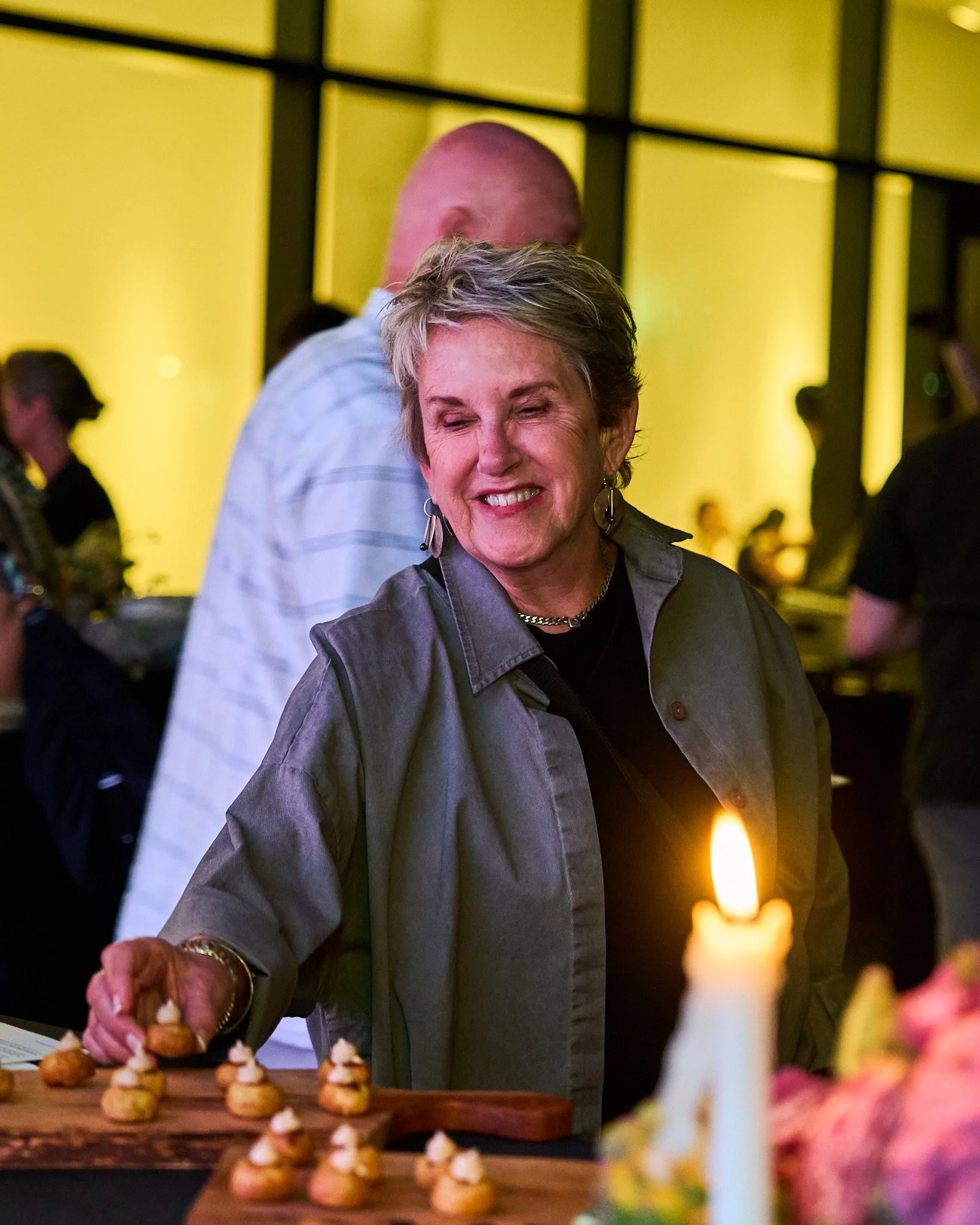 A woman with short gray hair, wearing earrings and a necklace, smiles while lighting a candle at a celebration or event. The background has a yellow wall with black frames and blurred people. Photographed by Gumption Studio, Ottawa event photographer