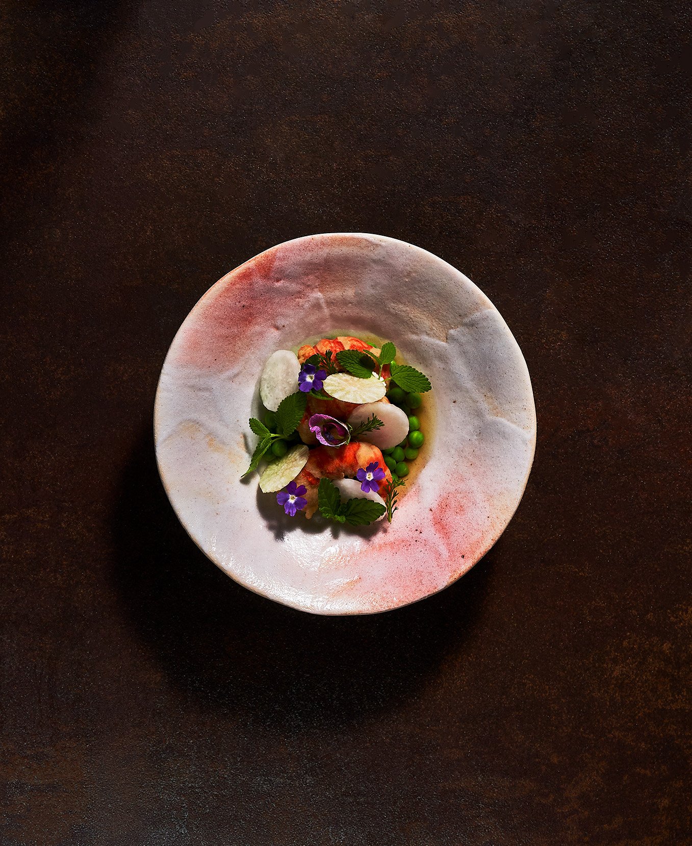 Peas and Prawns dish by Justin Chagampagne-Lagarde garnished with edible flowers, fresh herbs, and small green spheres, set against a dark background. Photographed for the Perch Cookbook by Gumption Studio, Ottawa Cookbook photographer.
