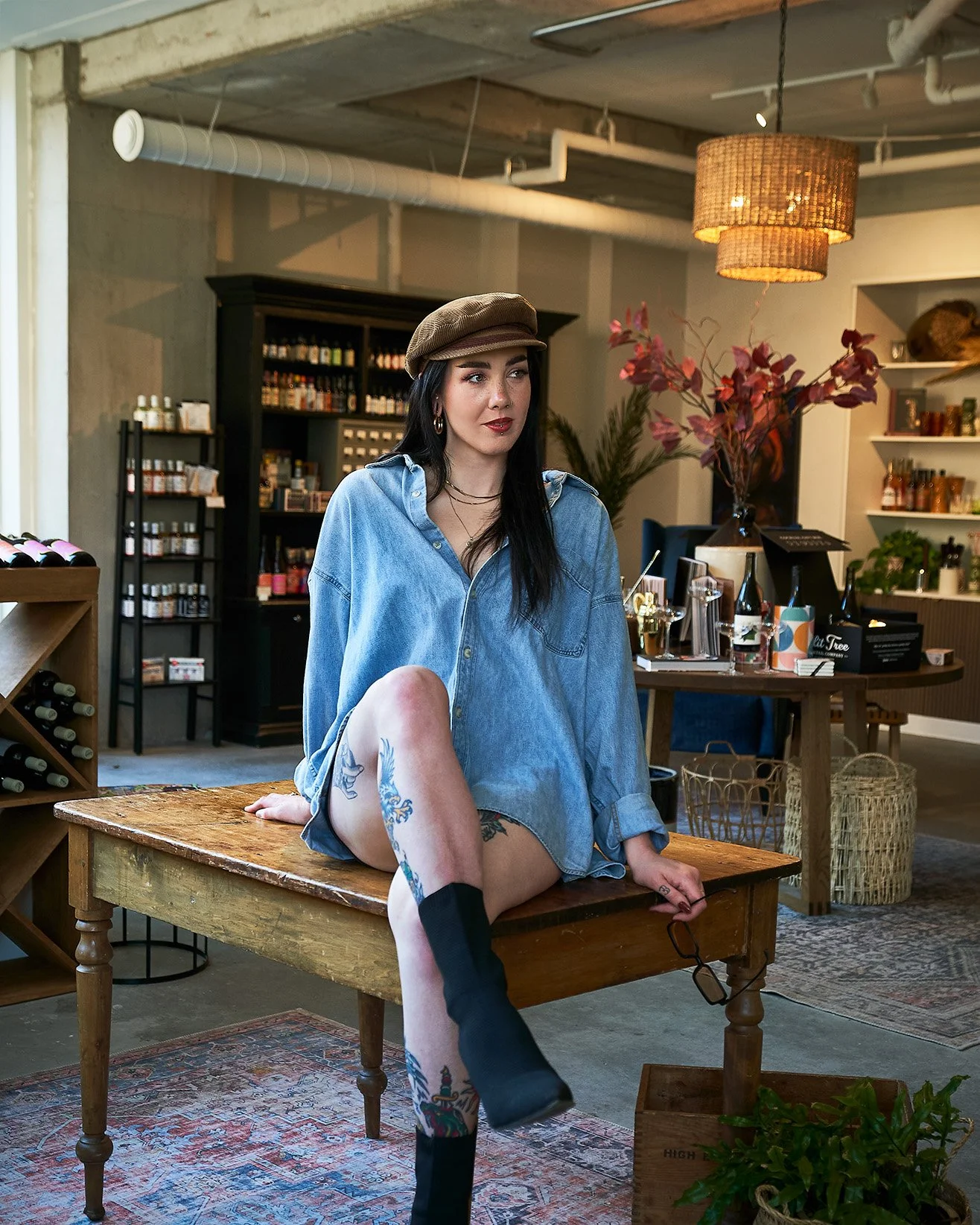 A woman with dark hair, tattoos, and pierced ears sits on a wooden table inside a stylish cafe or boutique. She is wearing a denim shirt, a brown cap, and black boots, with glasses hanging from her hand. The background features shelves with products,
