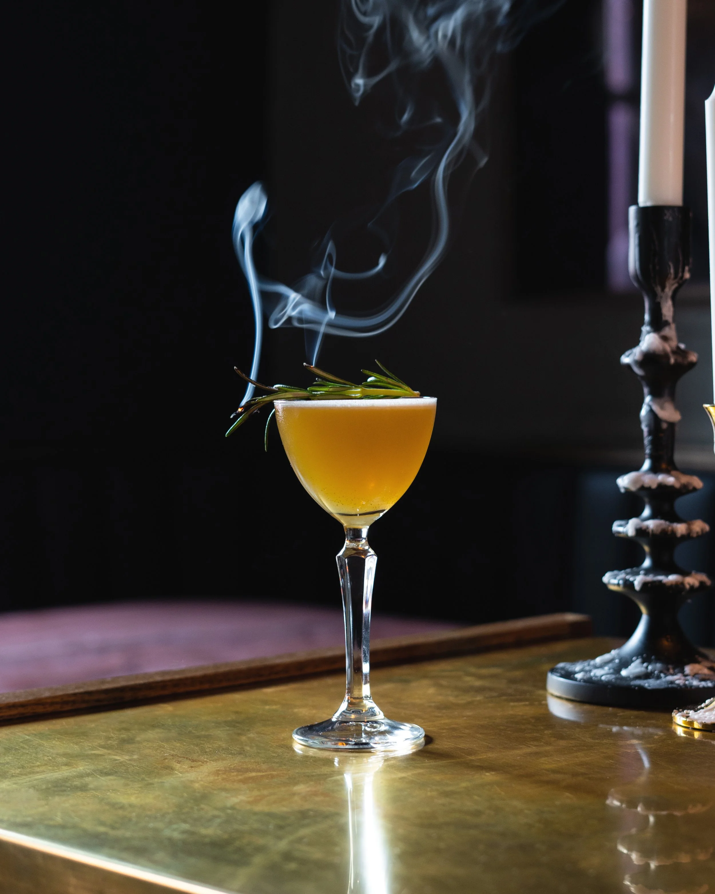 A cocktail drink in a tall, elegant glass garnished with a sprig of rosemary and emitting wisps of smoke or steam and a black candlestick with white wax melting on the right side. Photographed by Gumption Studio, Ottawa food photographer.