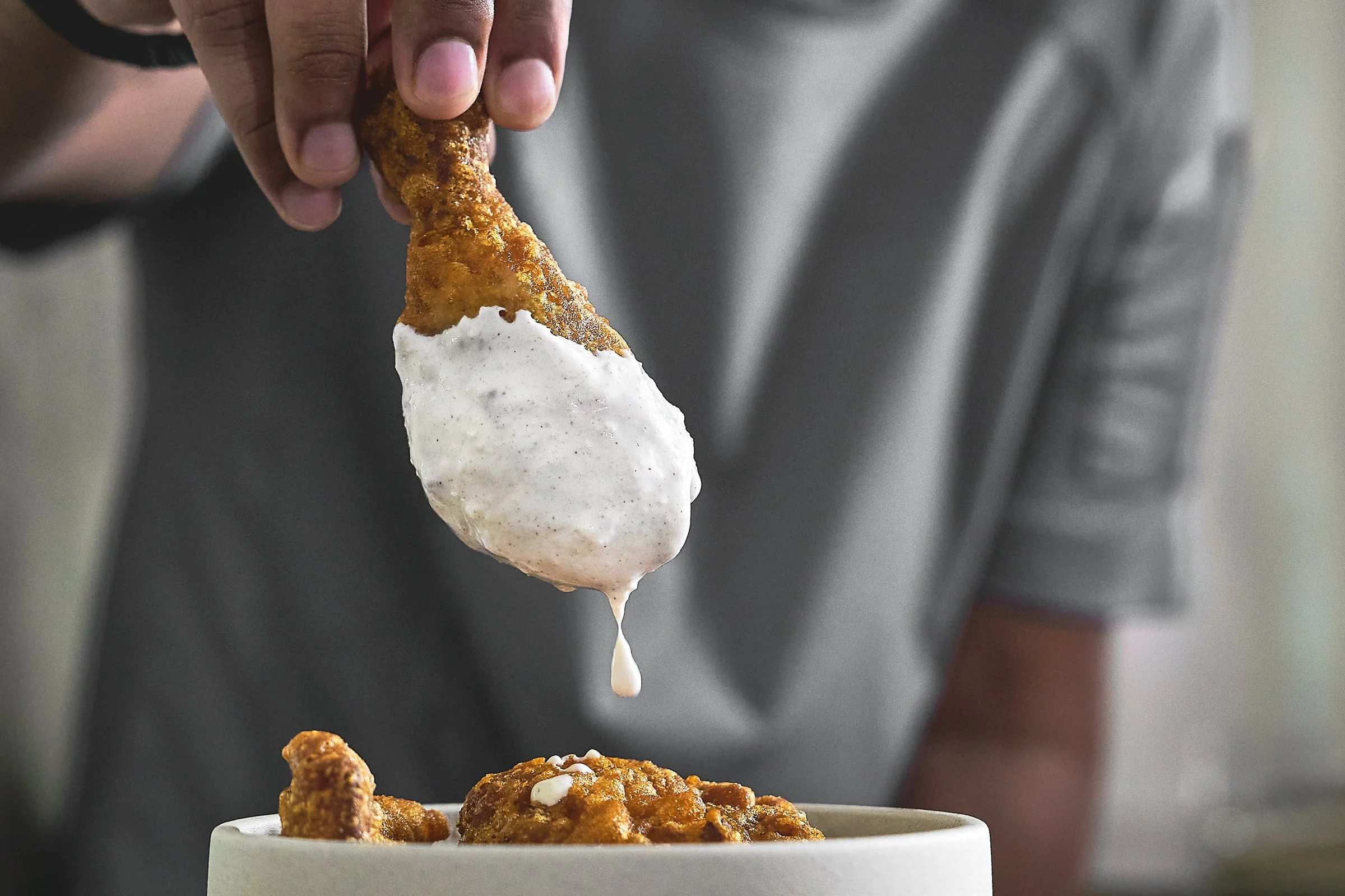 A close-up of a hand holding a fried chicken drumstick with white gravy dripping from it, over a bowl of chicken fried steak topped with white gravy. Kozhi Restaurant. Brand Identity