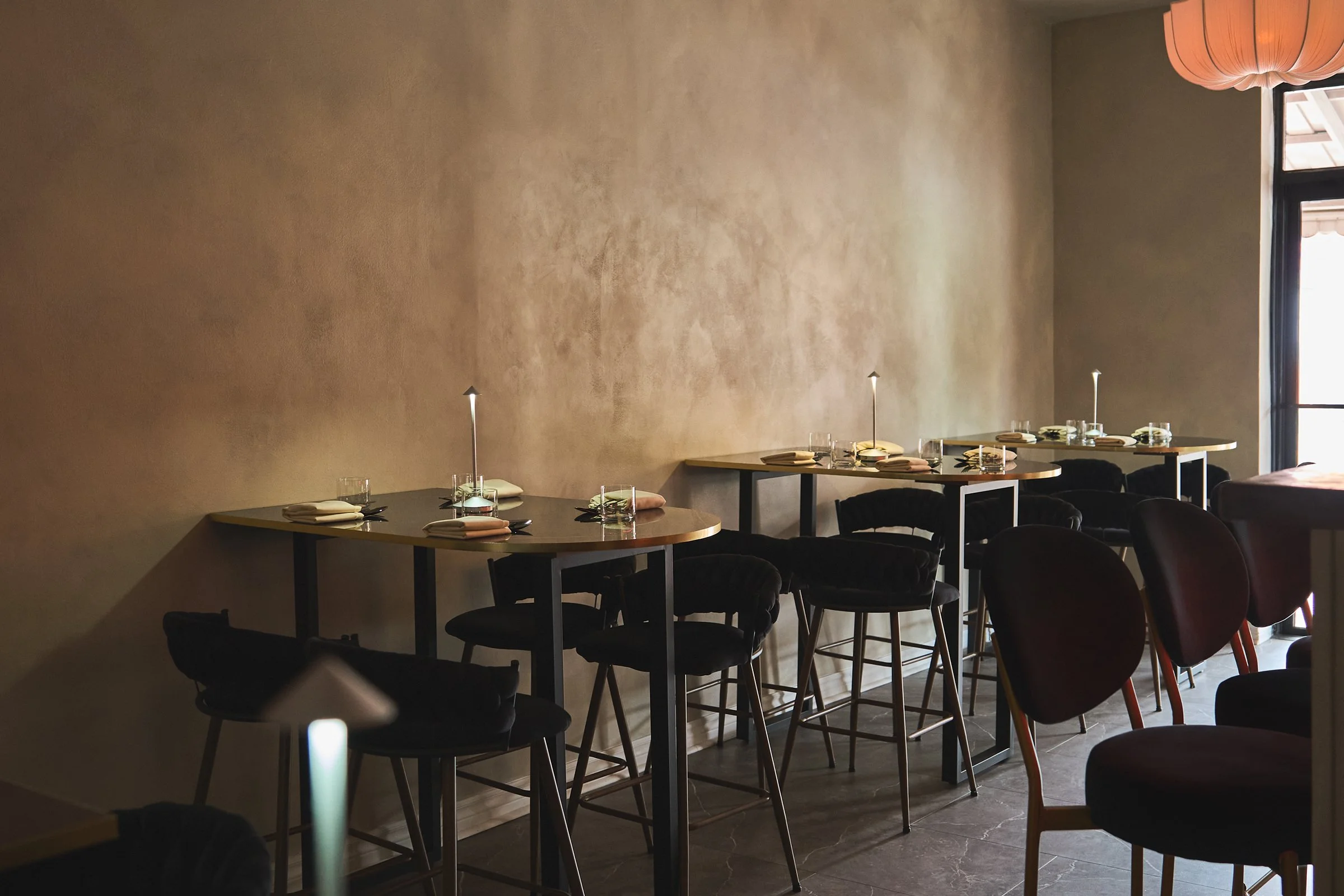 Empty restaurant with high tables set with plates, cutlery, napkins, and lamps, illuminated by natural light from windows. Kozhi Restaurant. Brand Identity