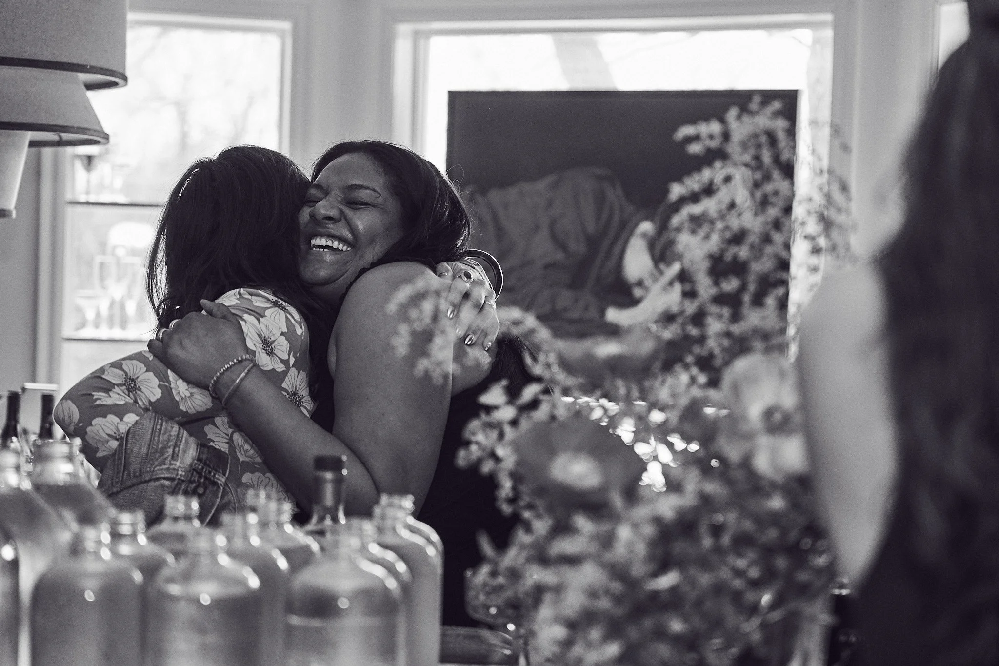 Two women hugging and smiling at a gathering, with a table of empty glasses and a painting in the background. Photographed by Gumption Studio, Ottawa event photographer.