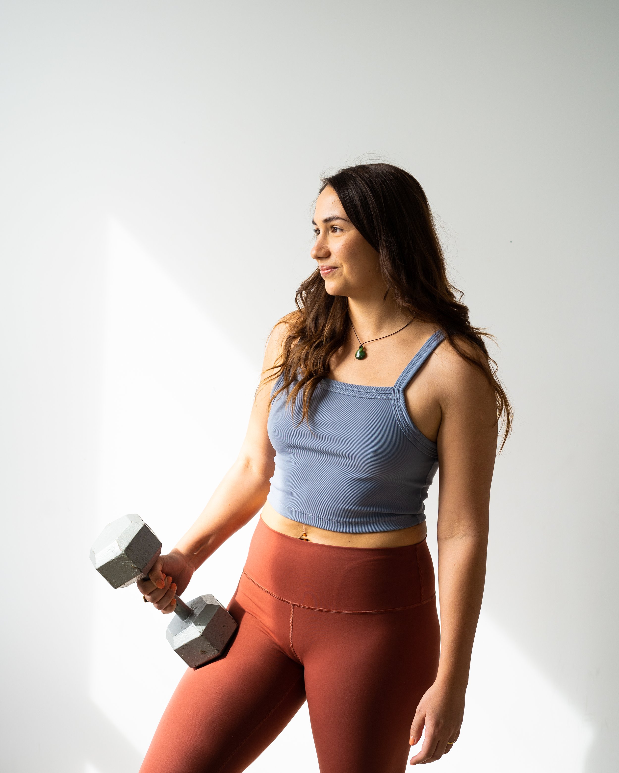 A woman with long dark hair wearing a blue sports bra and rust-colored leggings, holding a gray dumbbell in her right hand, standing against a plain white background.