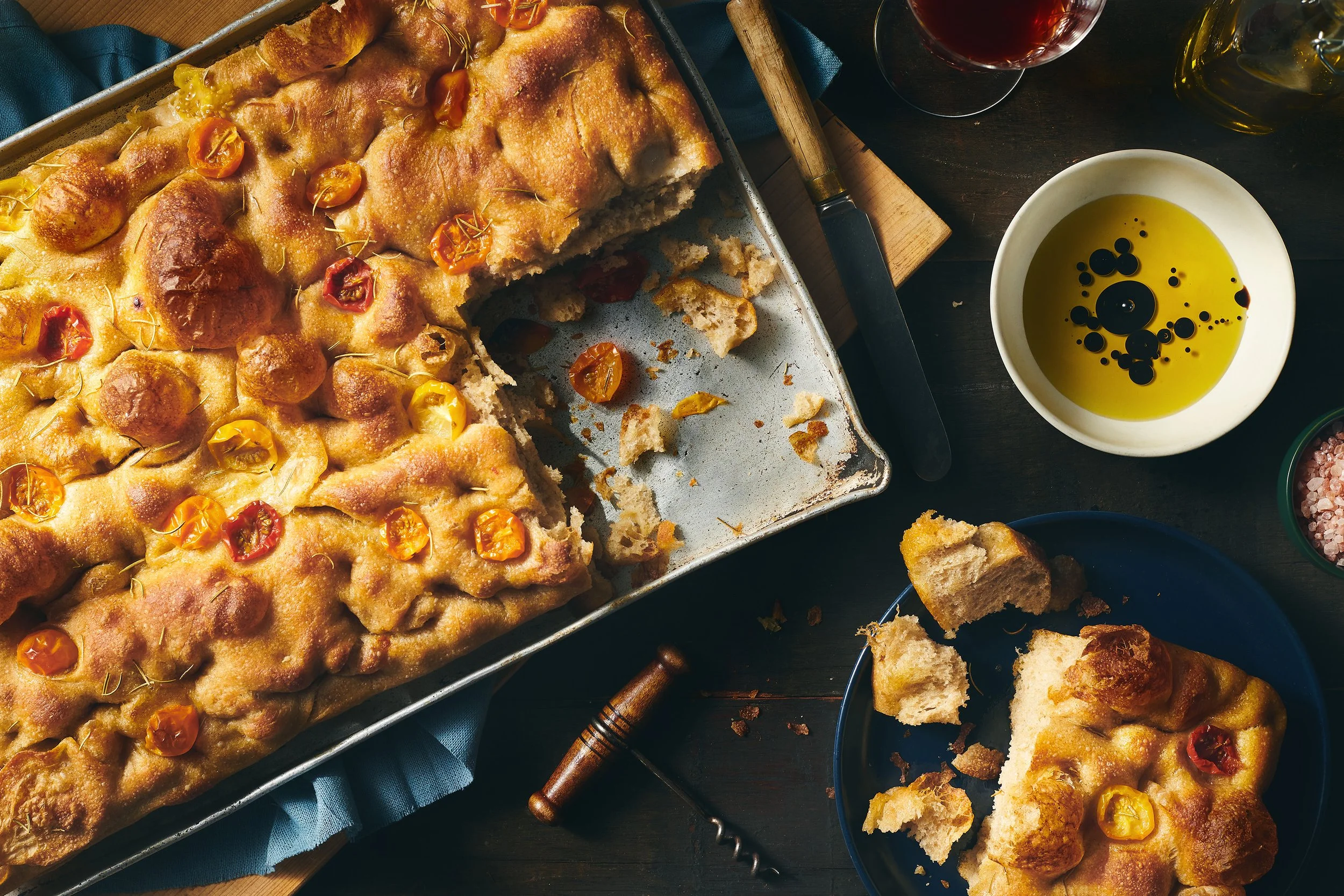 'Your Bread Box' focaccia bread topped with cherry tomatoes and herbs, partially sliced on a baking sheet. Surrounding the bread are a bowl of olive oil with balsamic vinegar. Photographed by Nick Ghattas, Ottawa food stylist is Gumption Studio.