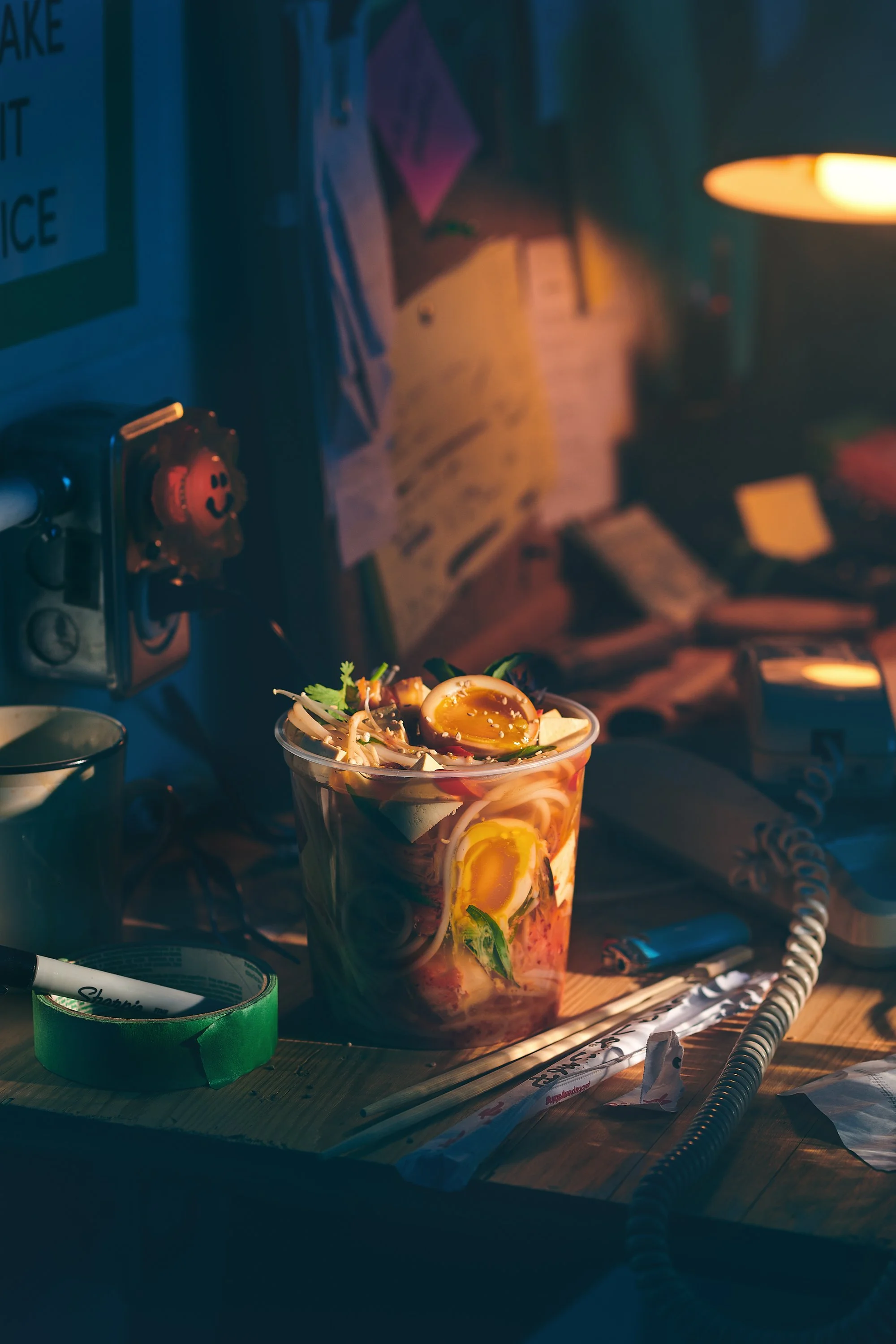 A plastic cup filled with a ramen and a sliced boiled eggs, greens, and other vegetables, placed on a wooden table illuminated by warm lighting. Photographed by Nick Ghattas, and Ottawa food stylist Gumption Studio.