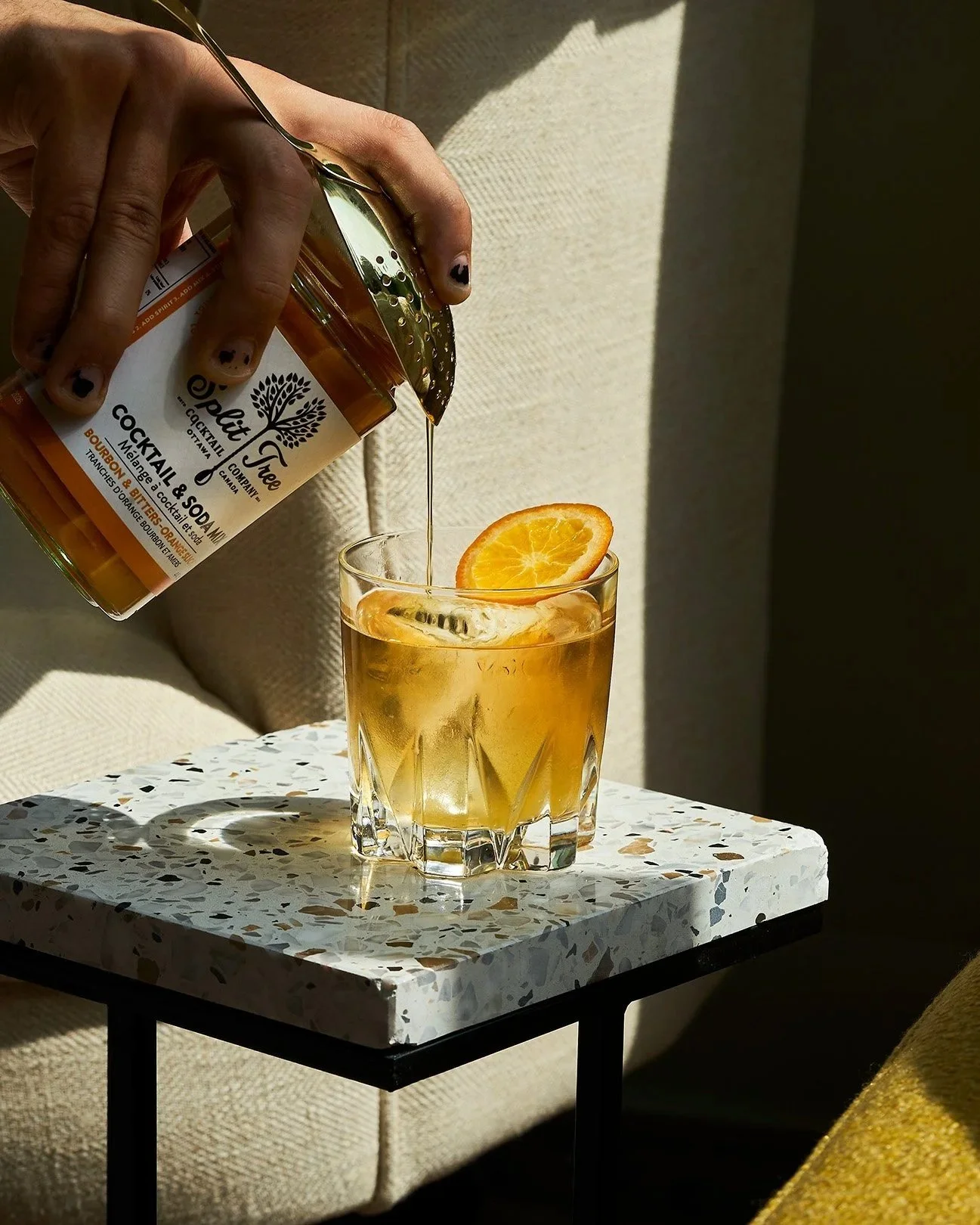 A hand pouring a Split Tree syrup into a glass with ice and an orange slice garnish on a small terrazzo table, with a beige cushioned chair and shadows in the background. Photographed by Gumption Studio, Ottawa cocktail photographer.