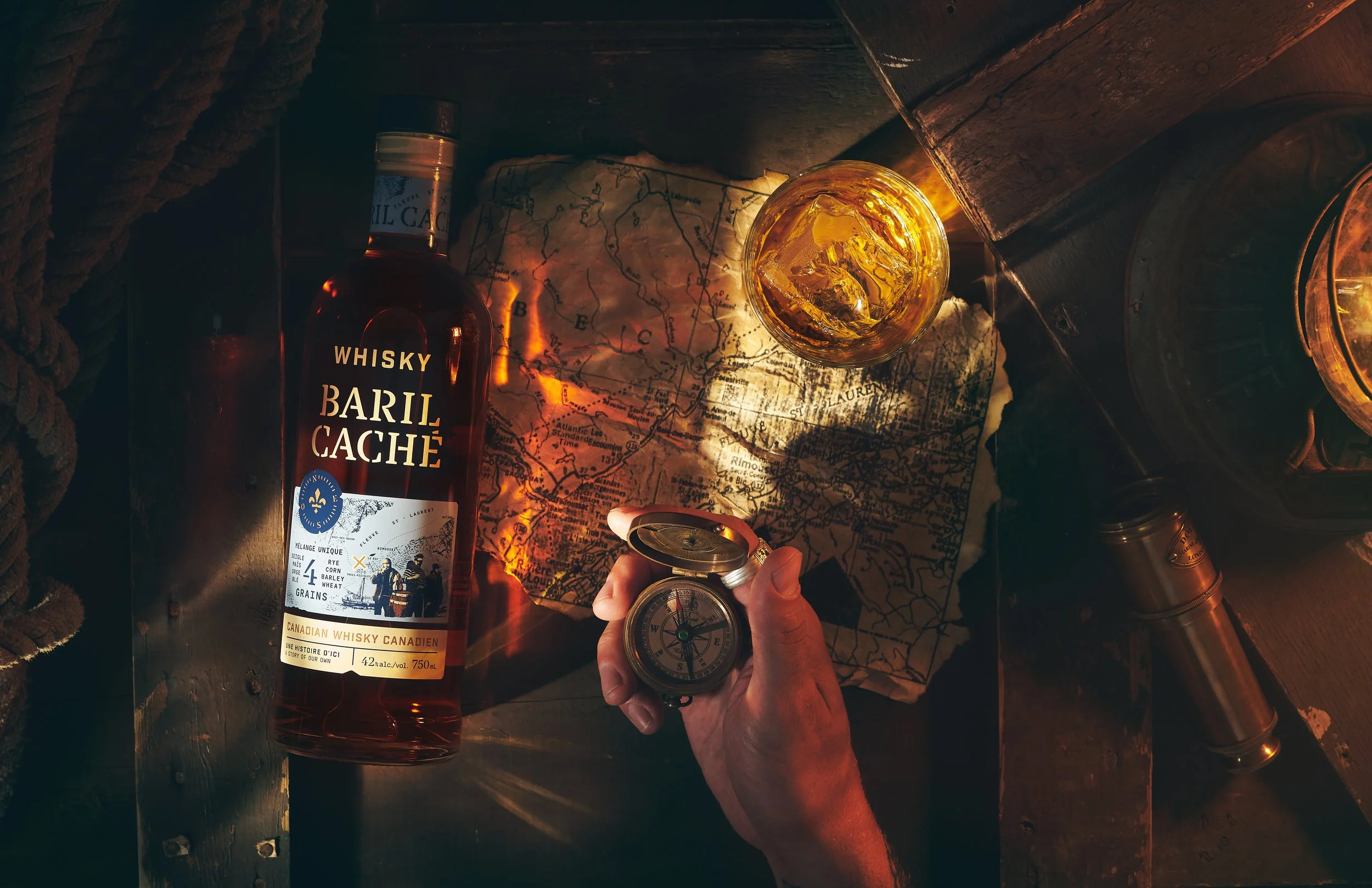 A bottle of 'Baril Caché' whiskey, a glass of whiskey with ice, a map, a compass, and a lantern on a wooden surface, suggesting an adventure or exploration theme. Photographed by Nick Ghattas, Ottawa food stylist is Gumption Studio.