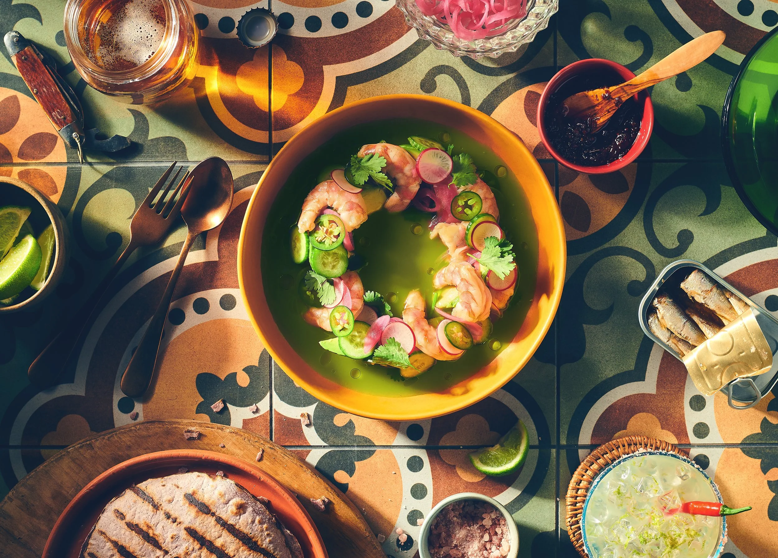 Colorful table setting with a bowl of shrimp and sliced radishes garnished with cilantro, a glass of beer, and various small bowls of condiments and sauces. Photographed by Nick Ghattas, food stylist is Gumption Studio.