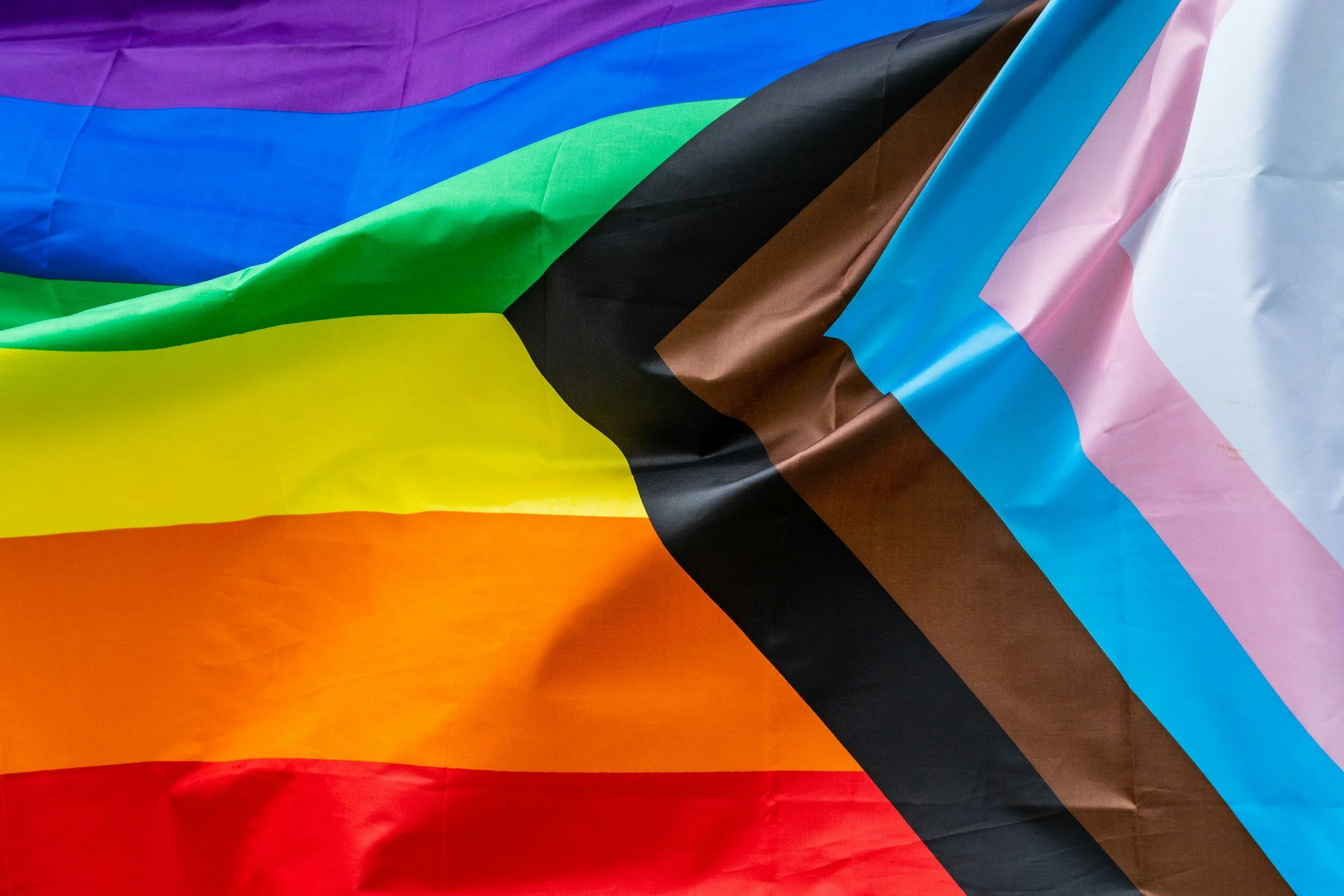 Close-up of a rainbow pride flag overlapping a transgender pride flag, both made of fabric with visible creases and folds.