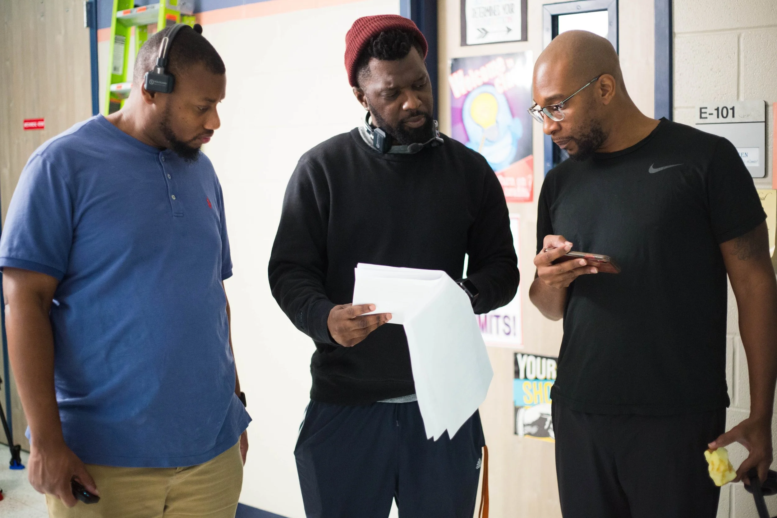 Three men standing together indoors, looking at a smartphone and a stack of paper. One man is wearing a blue polo shirt and headphones, another is in a black hoodie and red beanie, and the third is in a black t-shirt with glasses.