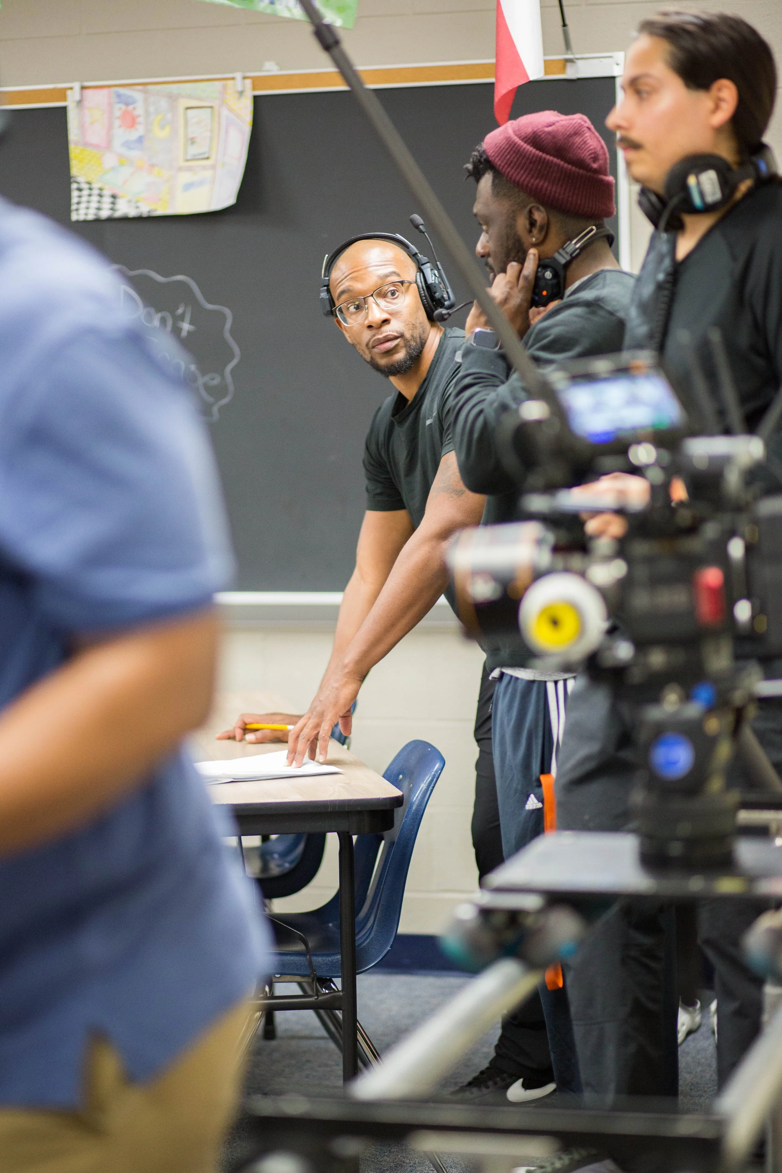 A group of people, including a man with glasses and headphones, sitting at a table in a classroom or studio setting, with a blackboard in the background and a camera in the foreground.