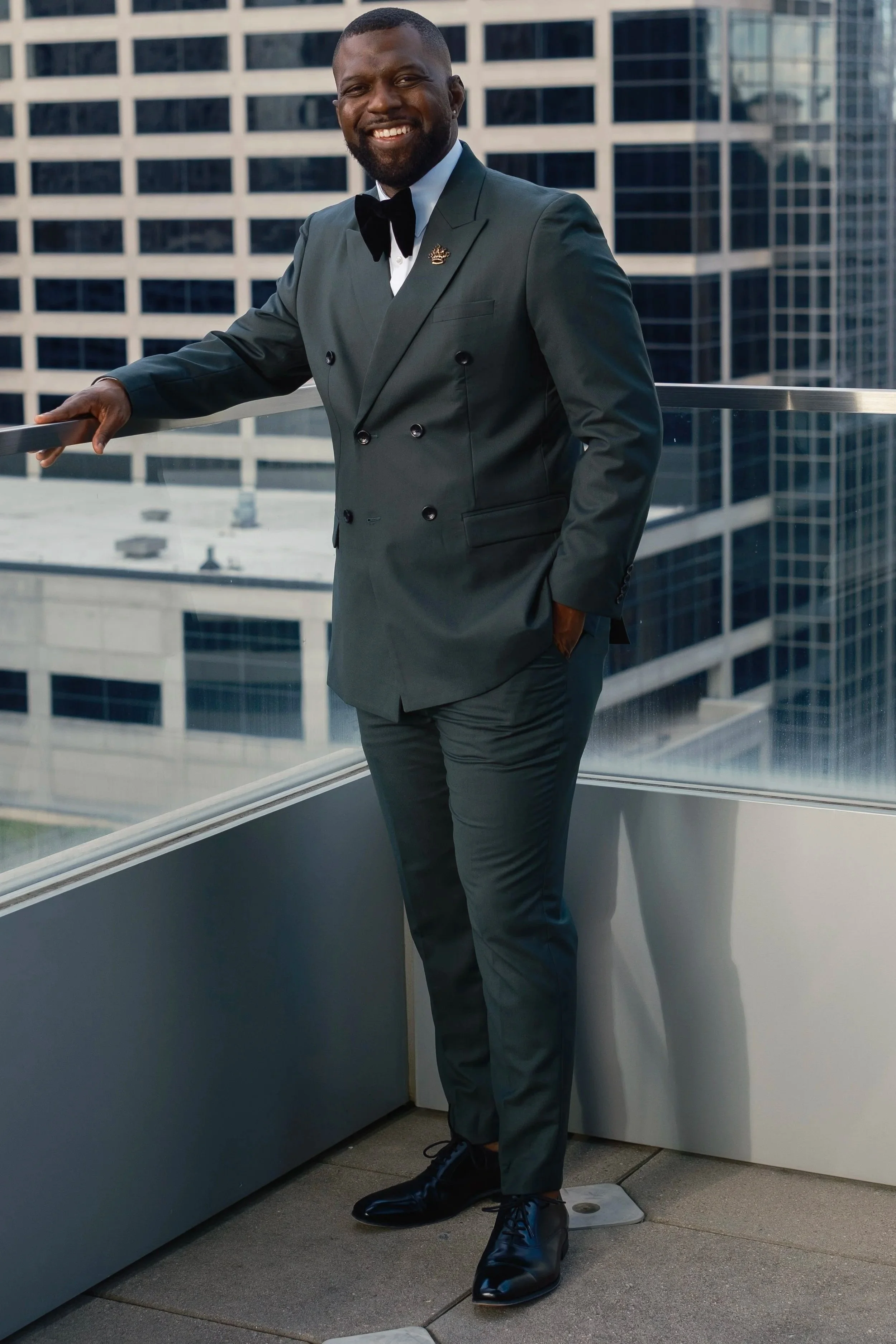 A man in a dark green double-breasted suit with a black bow tie, smiling and standing on a balcony with tall office buildings in the background.