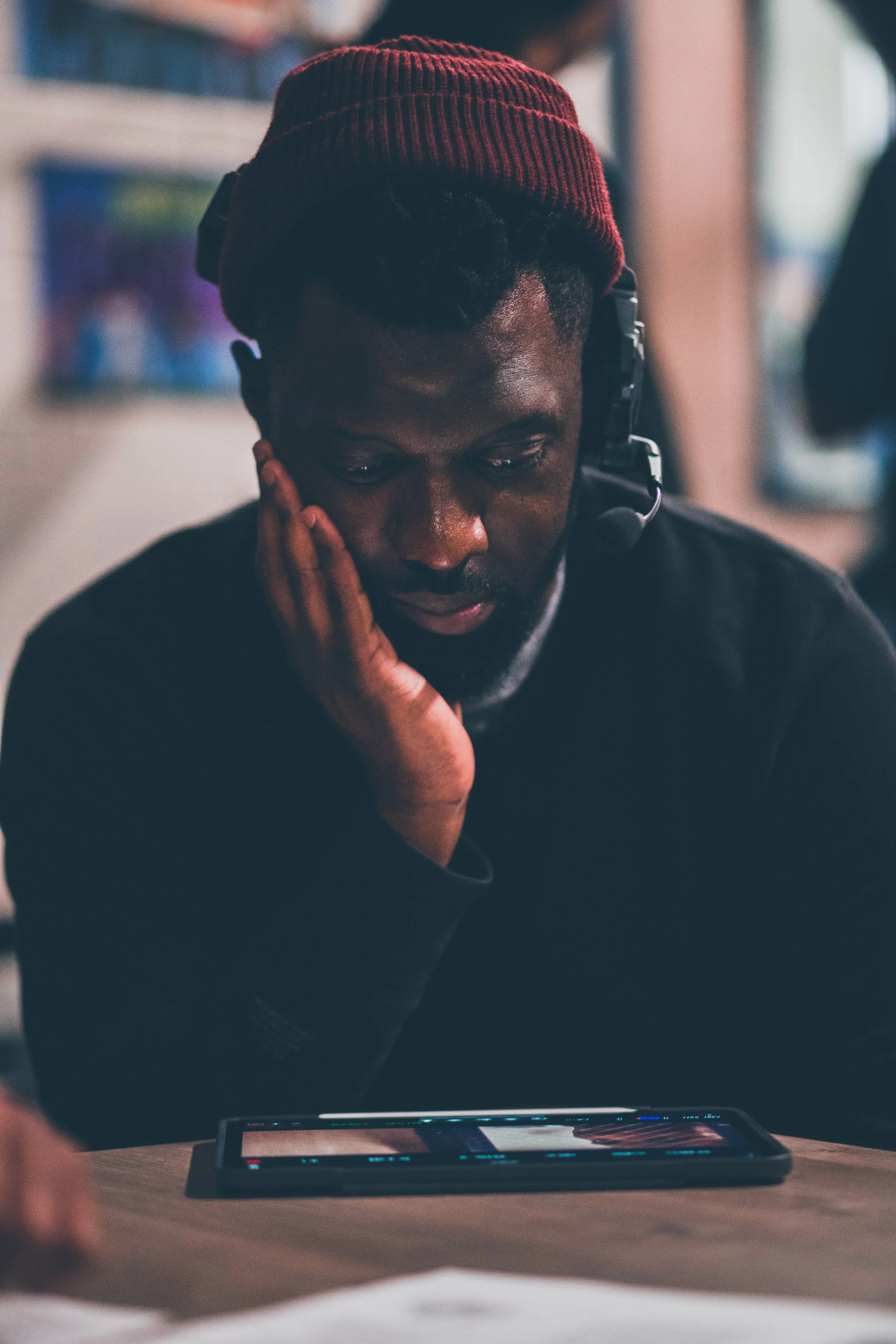 A man wearing a red beanie and black shirt, with a headset, sitting at a table and looking at a tablet with a focused expression.