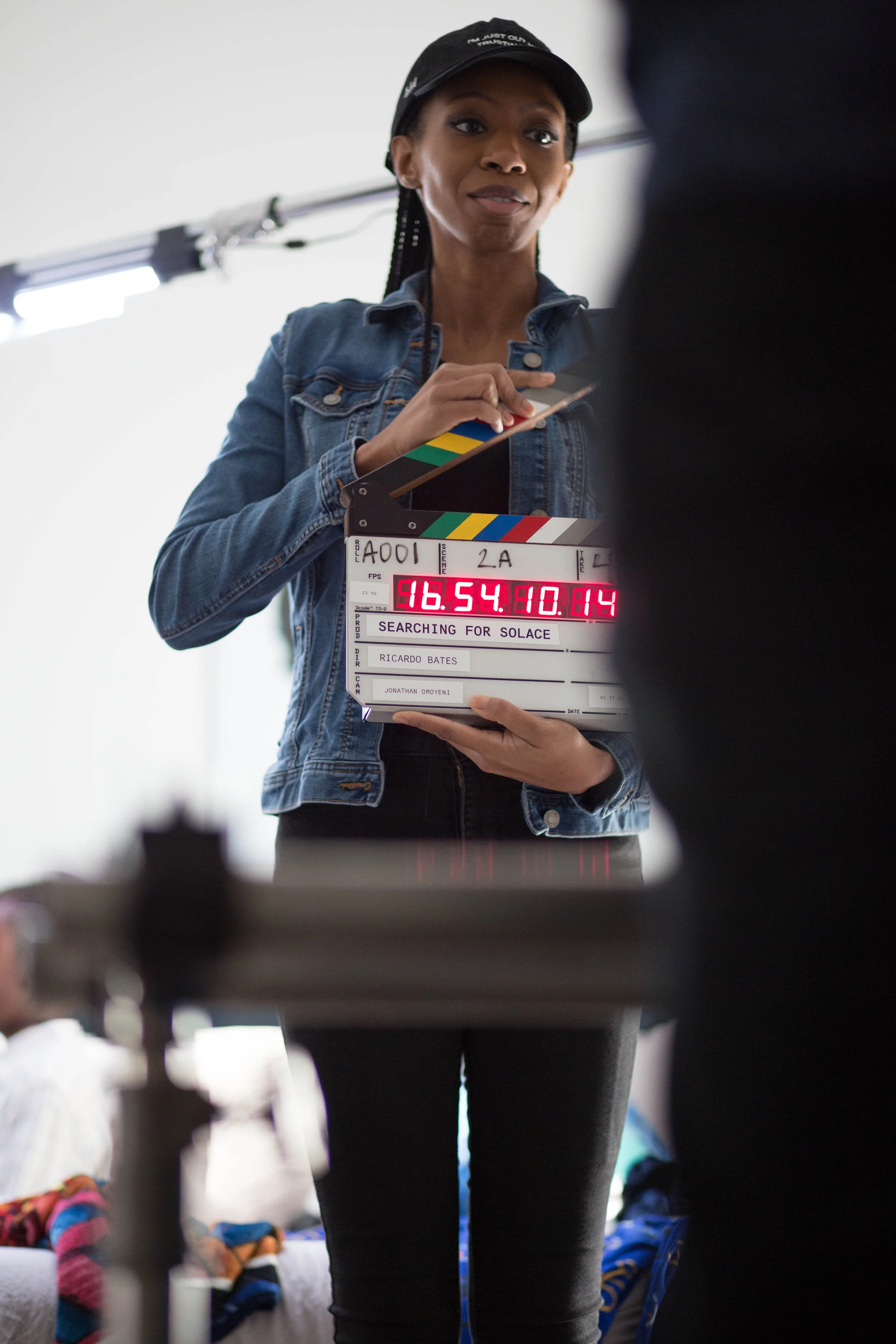 Clapperboard held by a woman wearing a denim jacket and black cap on film set.