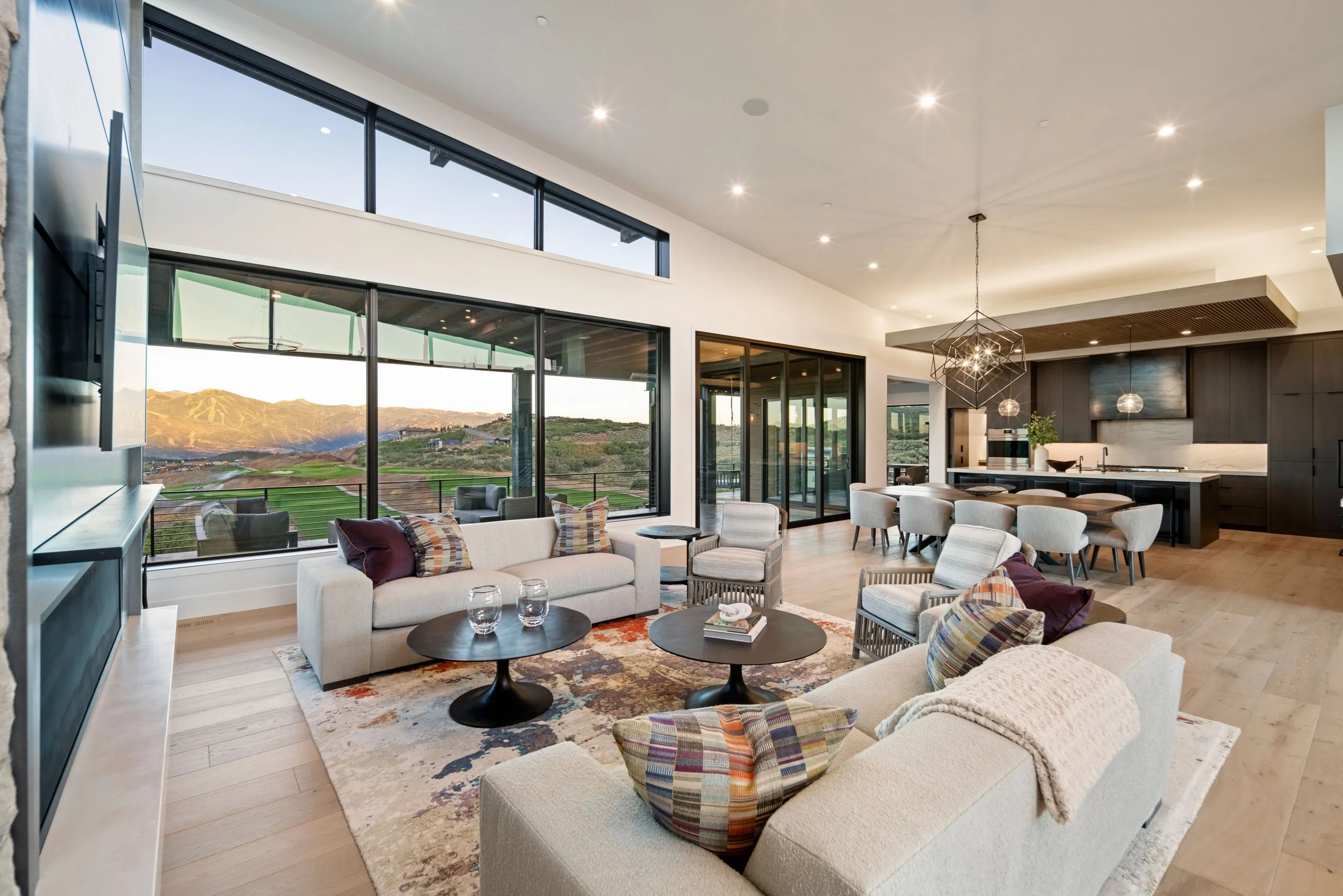 Spacious living room with white sofas, colorful pillows, wooden coffee tables, and large glass windows overlooking mountains and greenery, connected to an open dining area and modern kitchen.