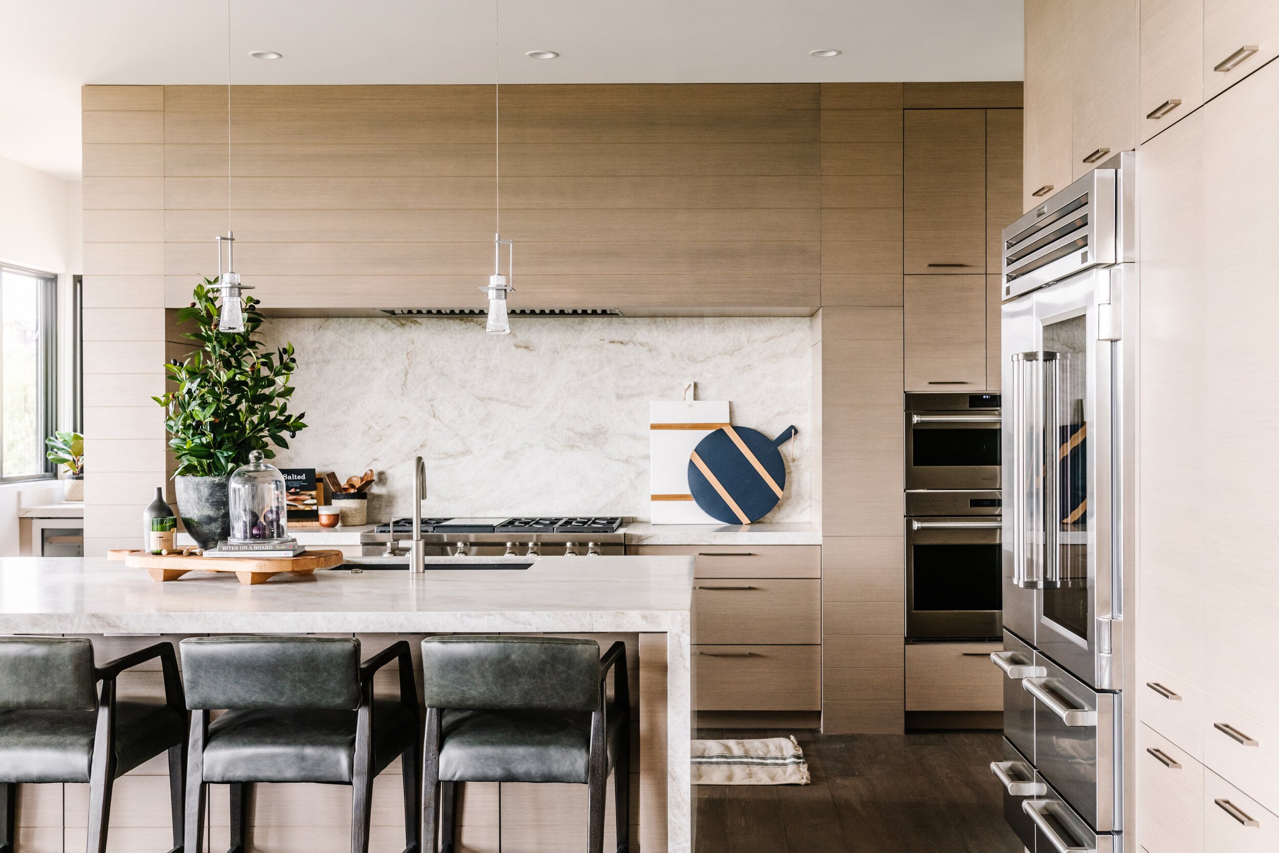 Modern kitchen with light wood cabinets, stainless steel appliances, a marble backsplash, a kitchen island with bar stools, and decorative items including a large potted plant and round cutting boards.