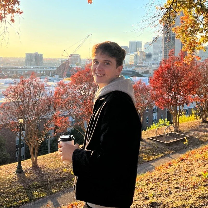 A young man in a black jacket with a hood, holding a coffee cup, smiling outdoors during sunset. The background features autumn trees with red and yellow leaves, a city skyline with tall buildings, and a construction crane.