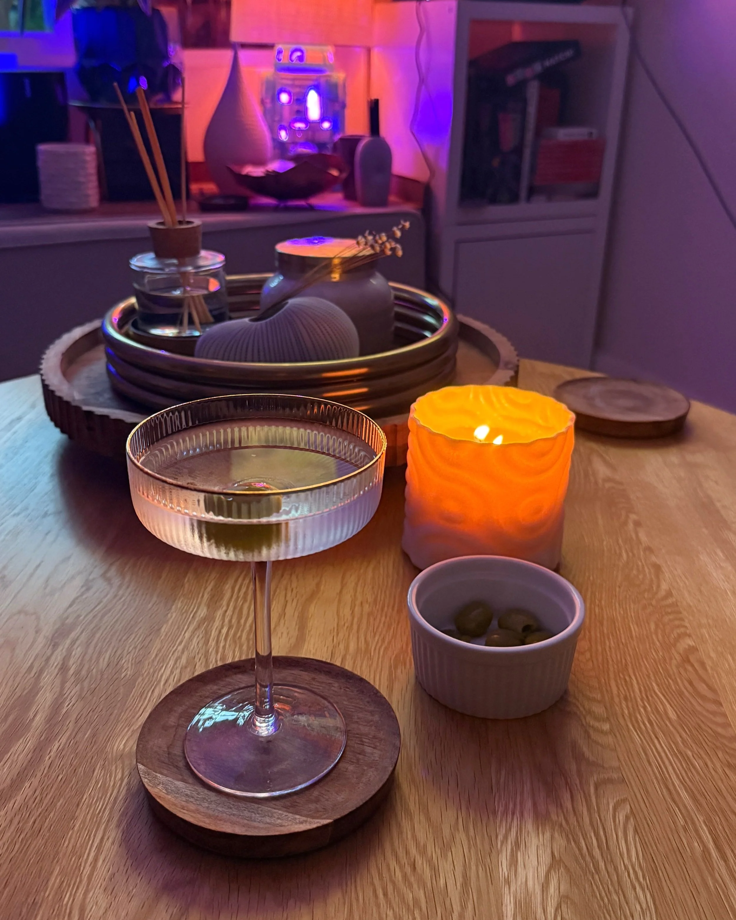 A wooden table with a cocktail in a coupe glass, a small bowl of green olives, a lit orange candle, and a tray of decorative vases and objects in the background with purple lighting.