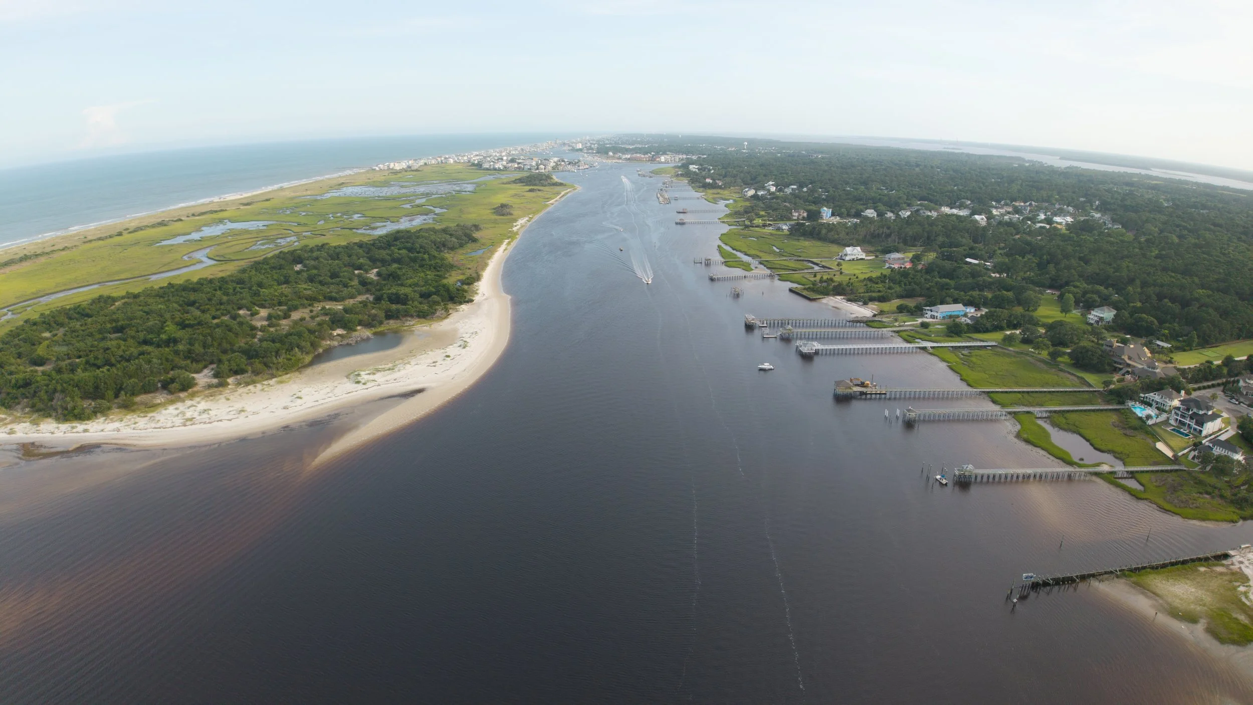 Aerial view of a coastal waterway with boats and docks, sandy beaches on the left, green marshland, and residential houses on the right.