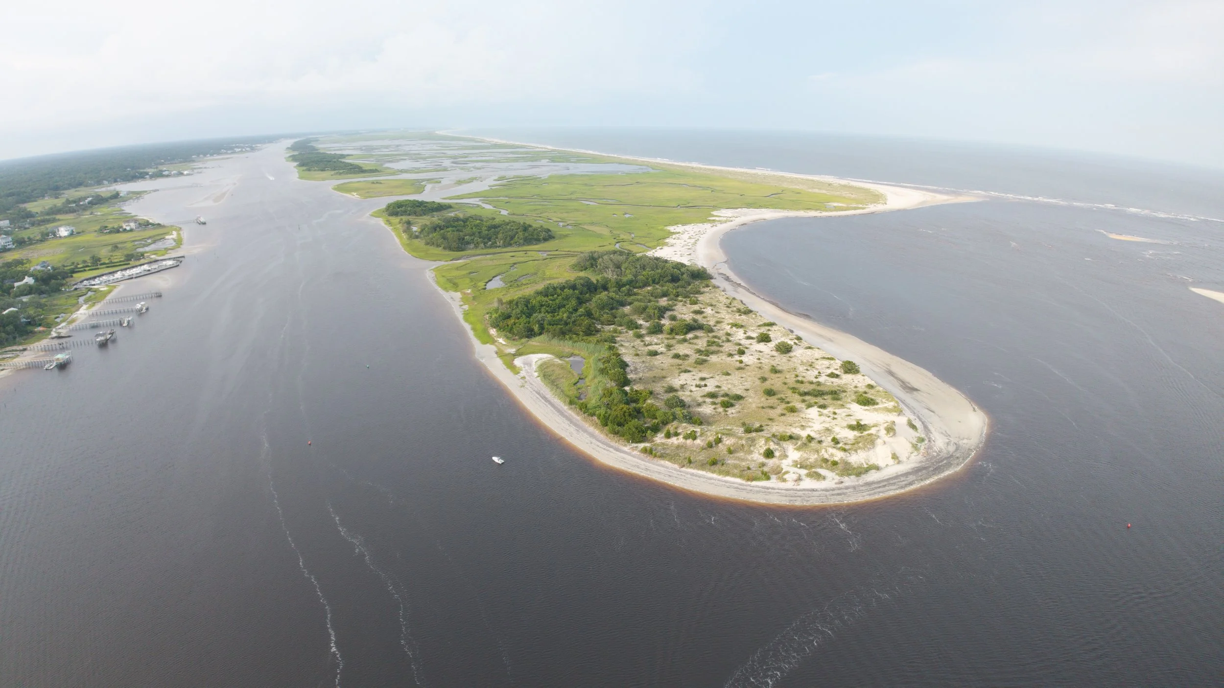 Aerial view of a coastal barrier island with sandy beaches, greenery, and surrounding water, with boats on the water and a cloudy sky overhead.