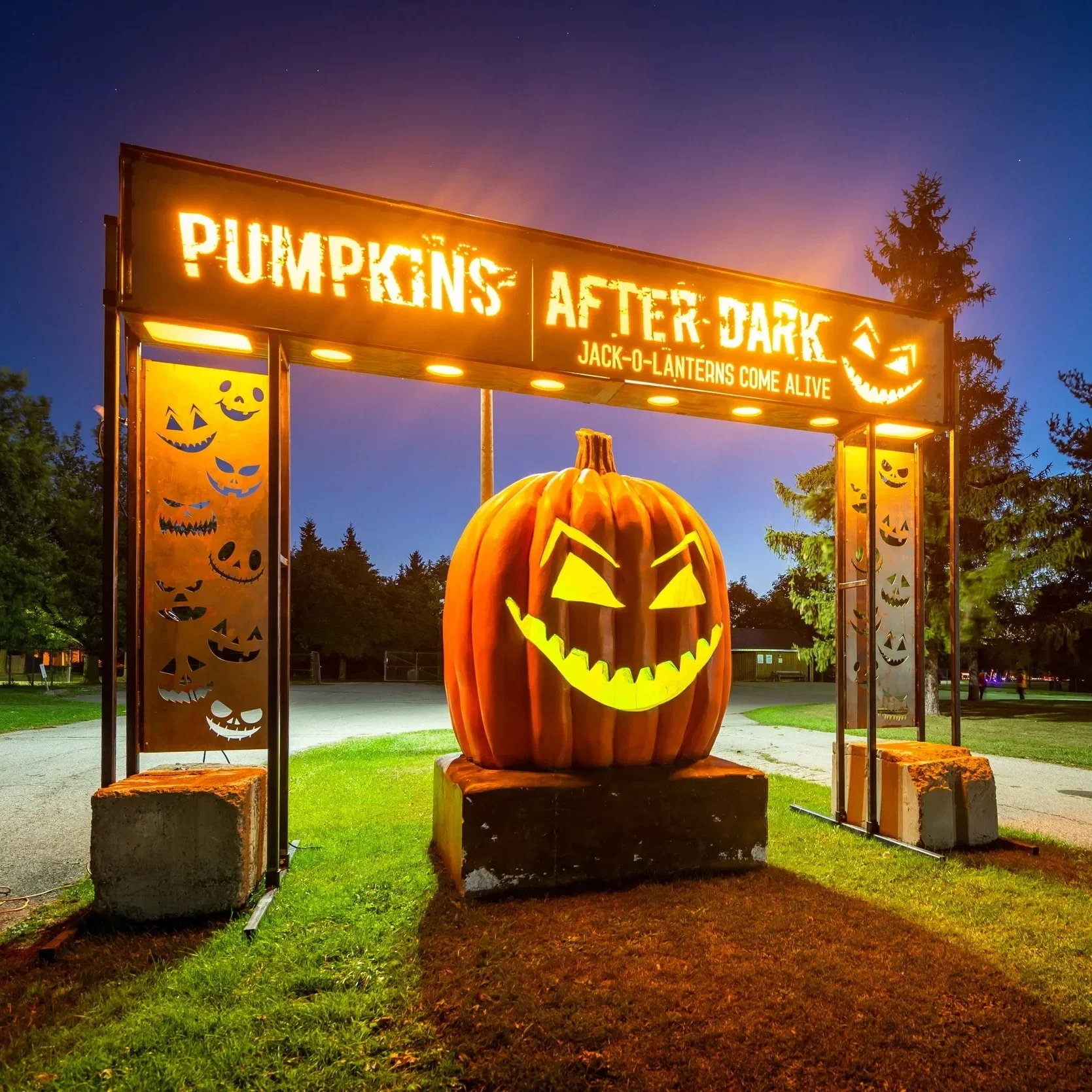Large illuminated pumpkin with a carved smiling face at night, surrounded by Halloween festive signage and decorations, in a park-like setting.