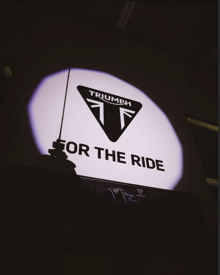 Projection of the Triumph logo and the text "FOR THE RIDE" on a wall in a dark room.