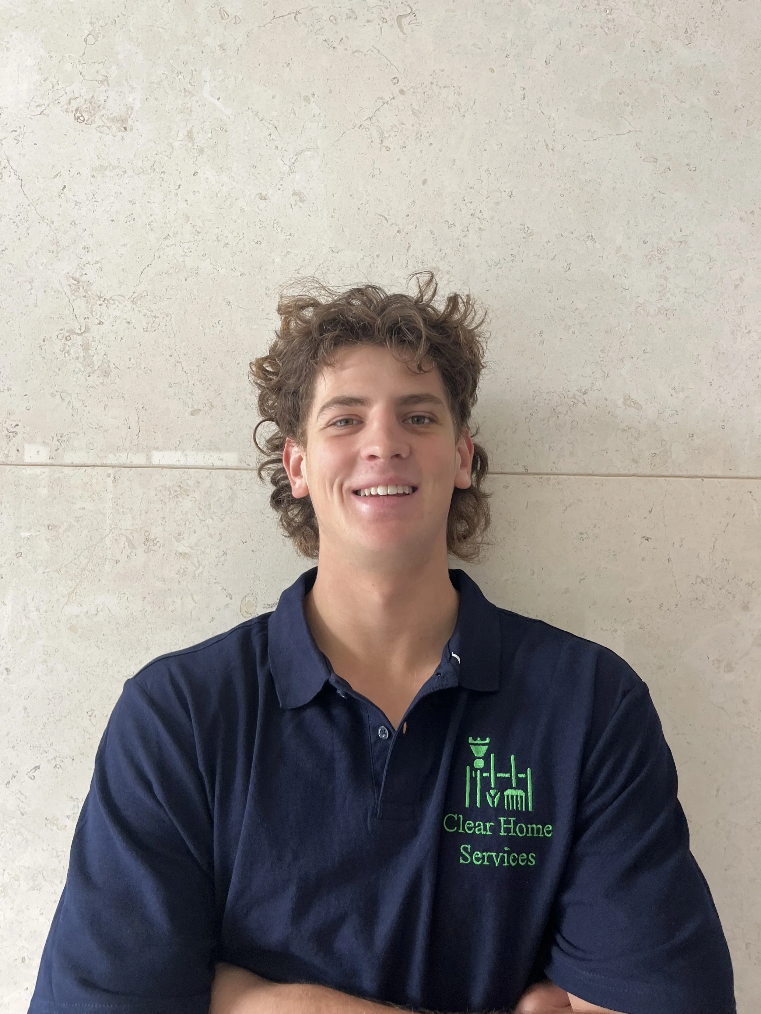Young man with curly hair wearing a navy blue polo shirt with a green logo and text that reads 'Clear Home Services,' standing against a beige tiled wall.