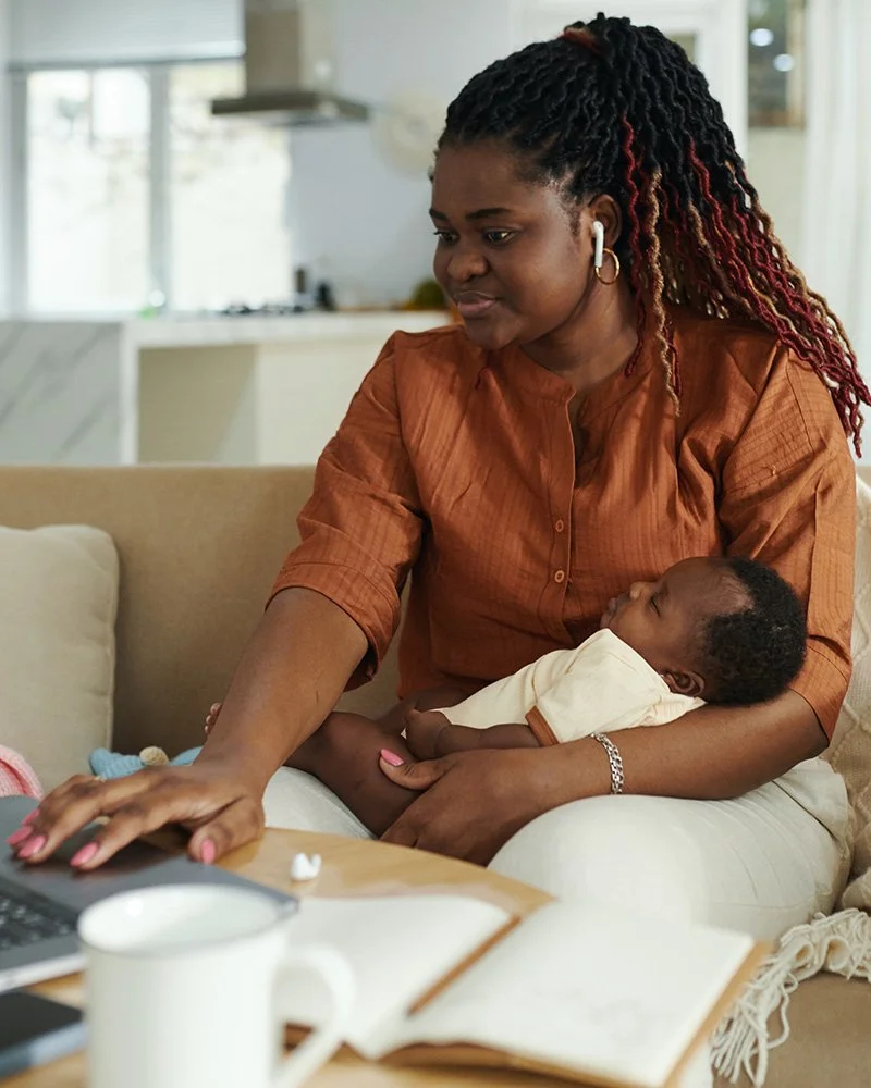 Parent sitting at a table with a laptop and coffee, holding a baby in their arms