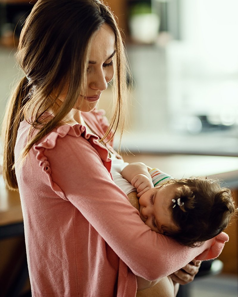 Smiling parent in a pink sweater holding a newborn while breastfeeding in a cozy home setting
