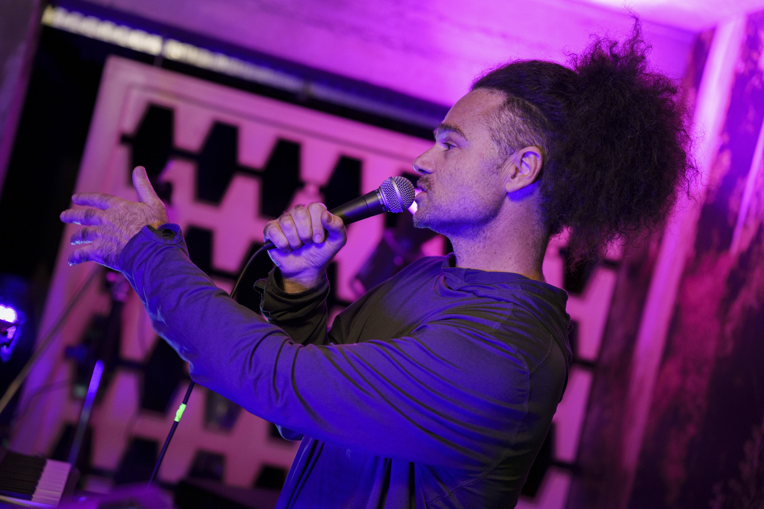 A person with long, curly hair, singing into a microphone, with purple and pink stage lighting in the background.