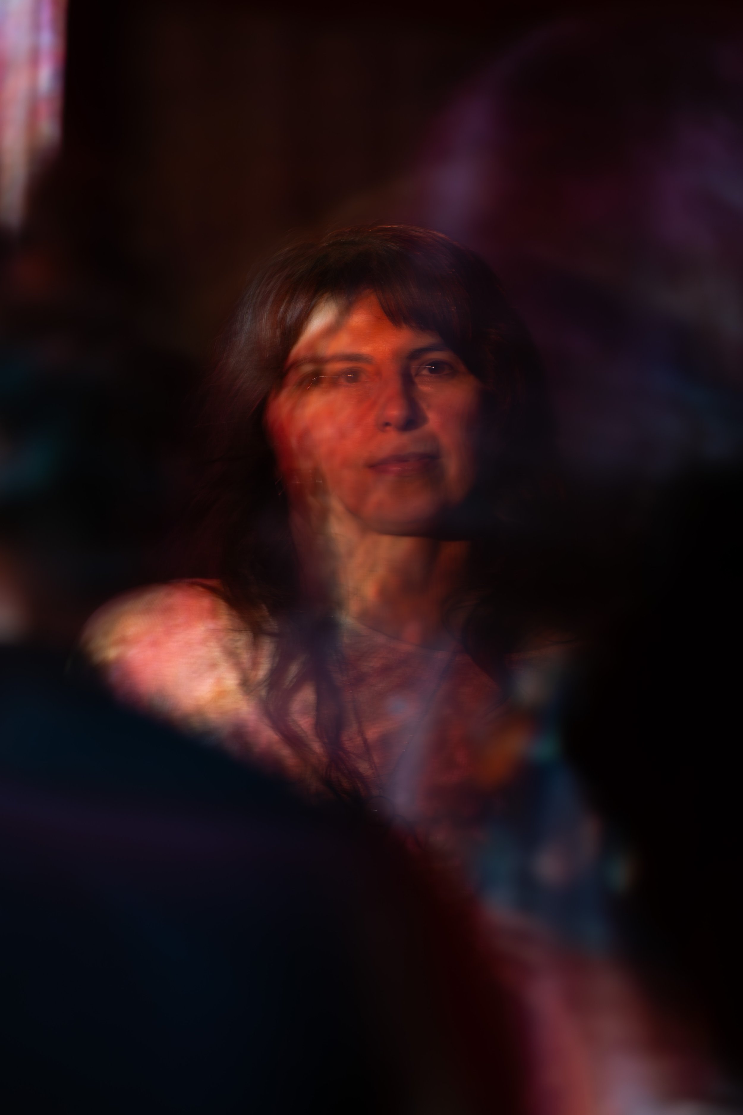 A woman with dark hair and a slight smile, illuminated by soft, colorful lighting with a blurred, dark background.