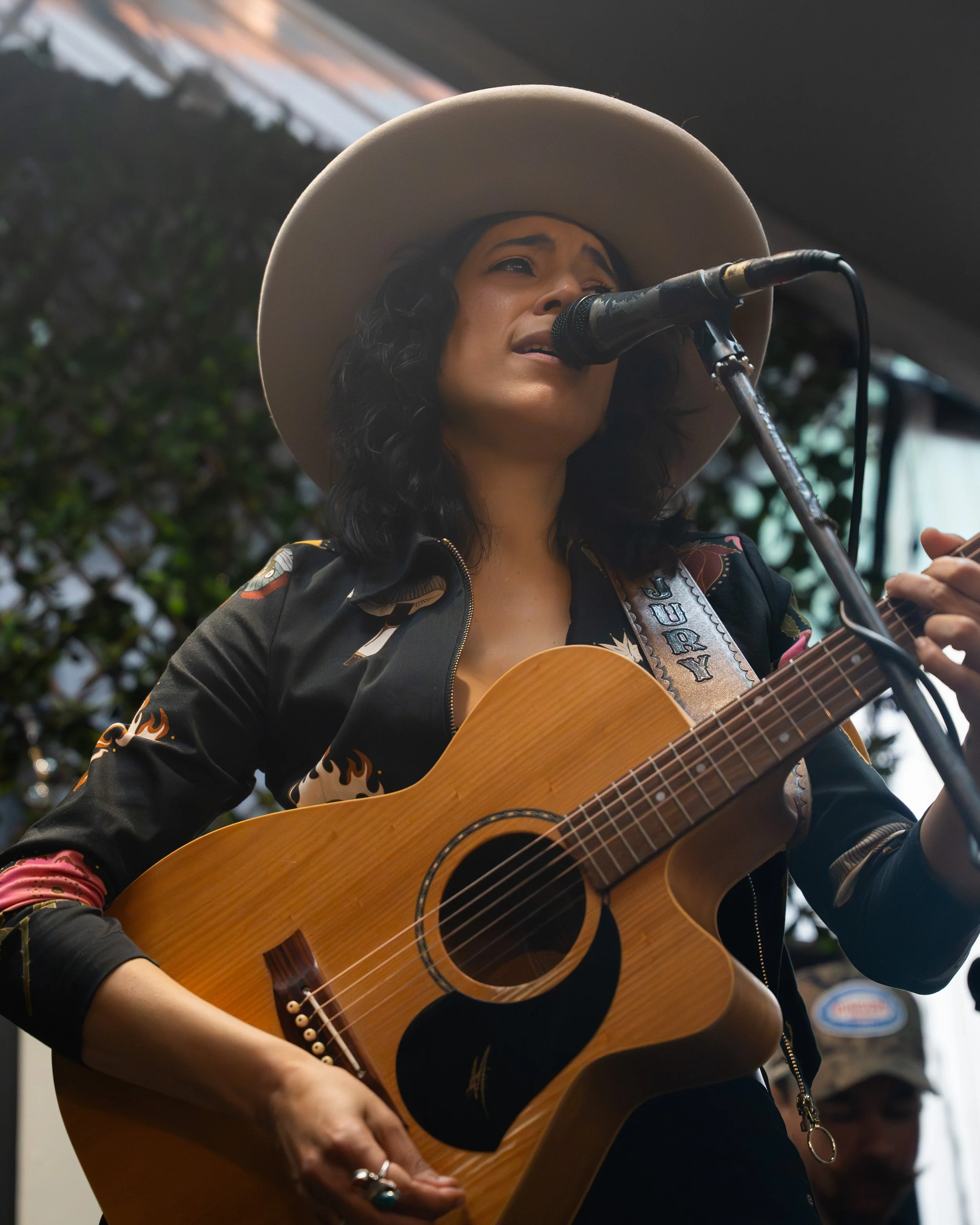 A woman singing passionately into a microphone while playing an acoustic guitar, wearing a large beige hat and a black jacket with colorful patterns, outdoors with greenery and some signs in the background.