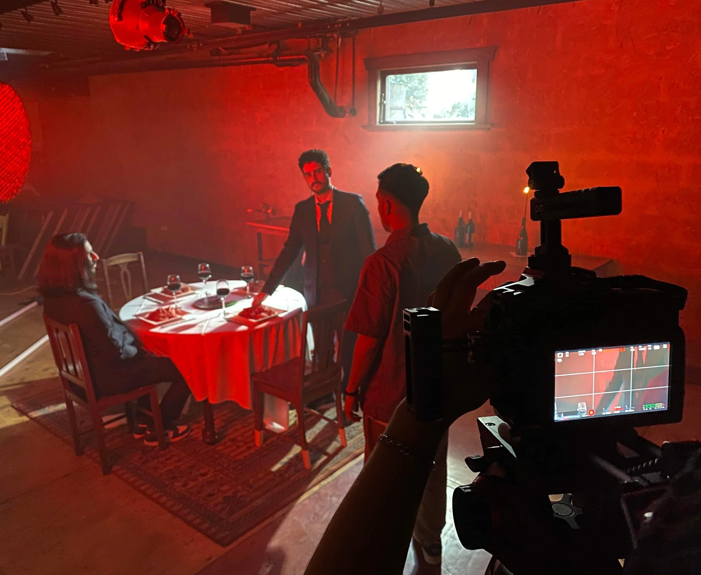 Behind-the-scenes of a music video production with three actors around a dining table and a camera operator filming the scene, all illuminated by red lighting.