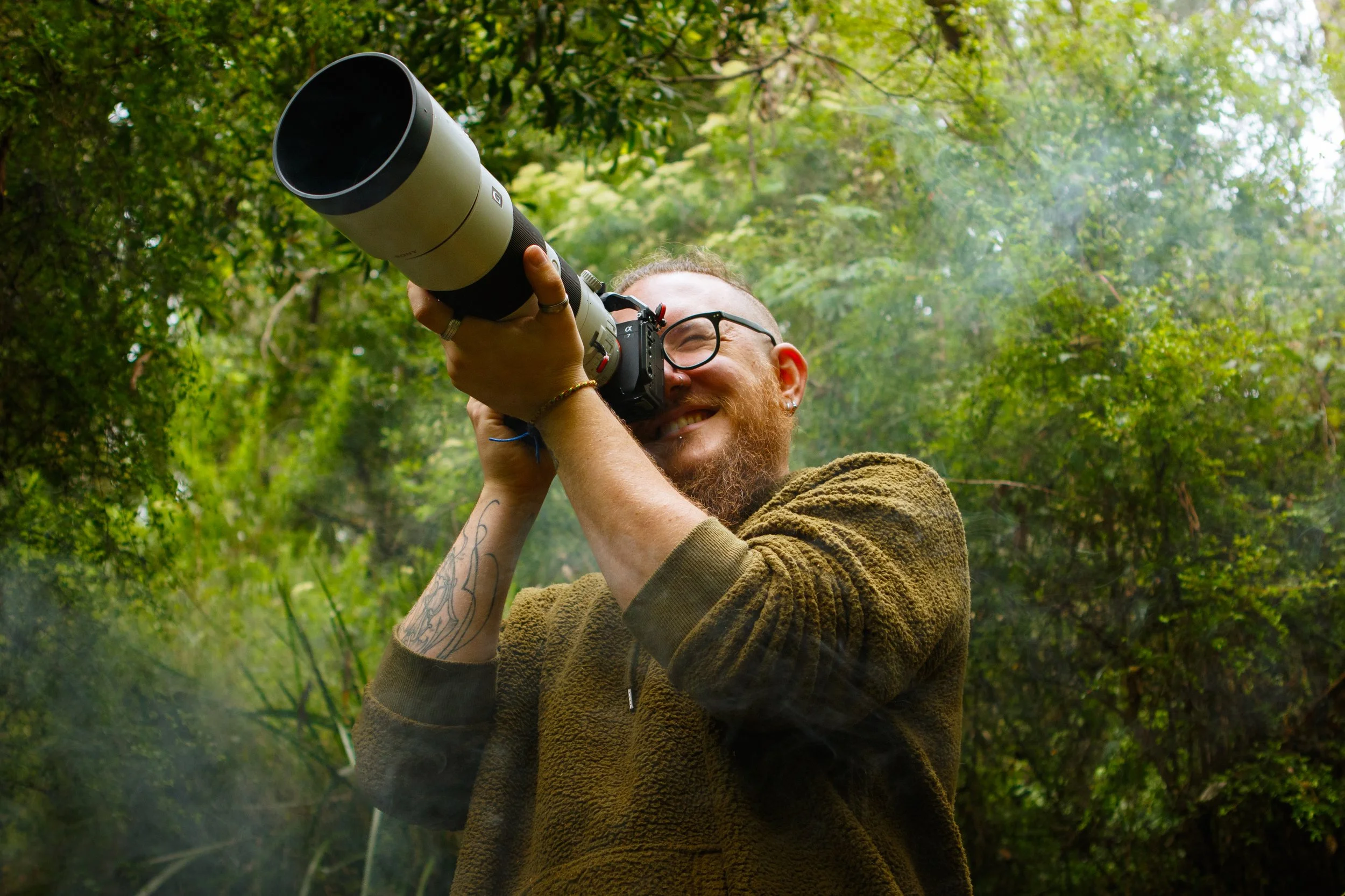A man with glasses and a beard smiling while taking a photograph outdoors in a green, wooded area, holding a camera with a large telephoto lens.
