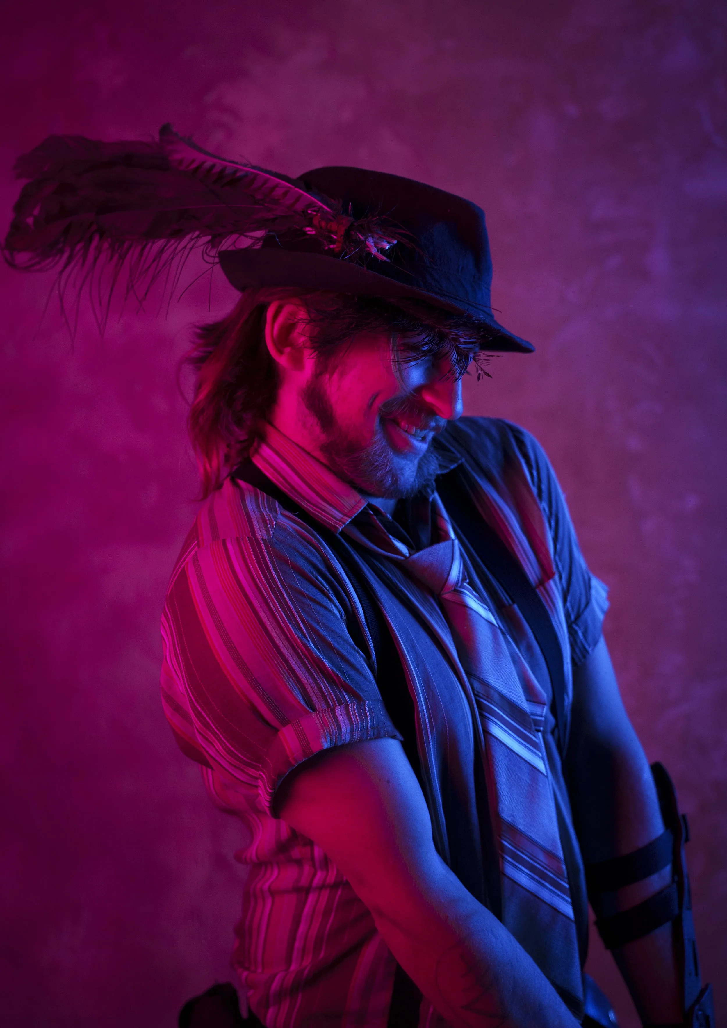 A man with long hair and a beard wearing a hat with feathers, illuminated by pink and blue lights, smiling and looking downward.
