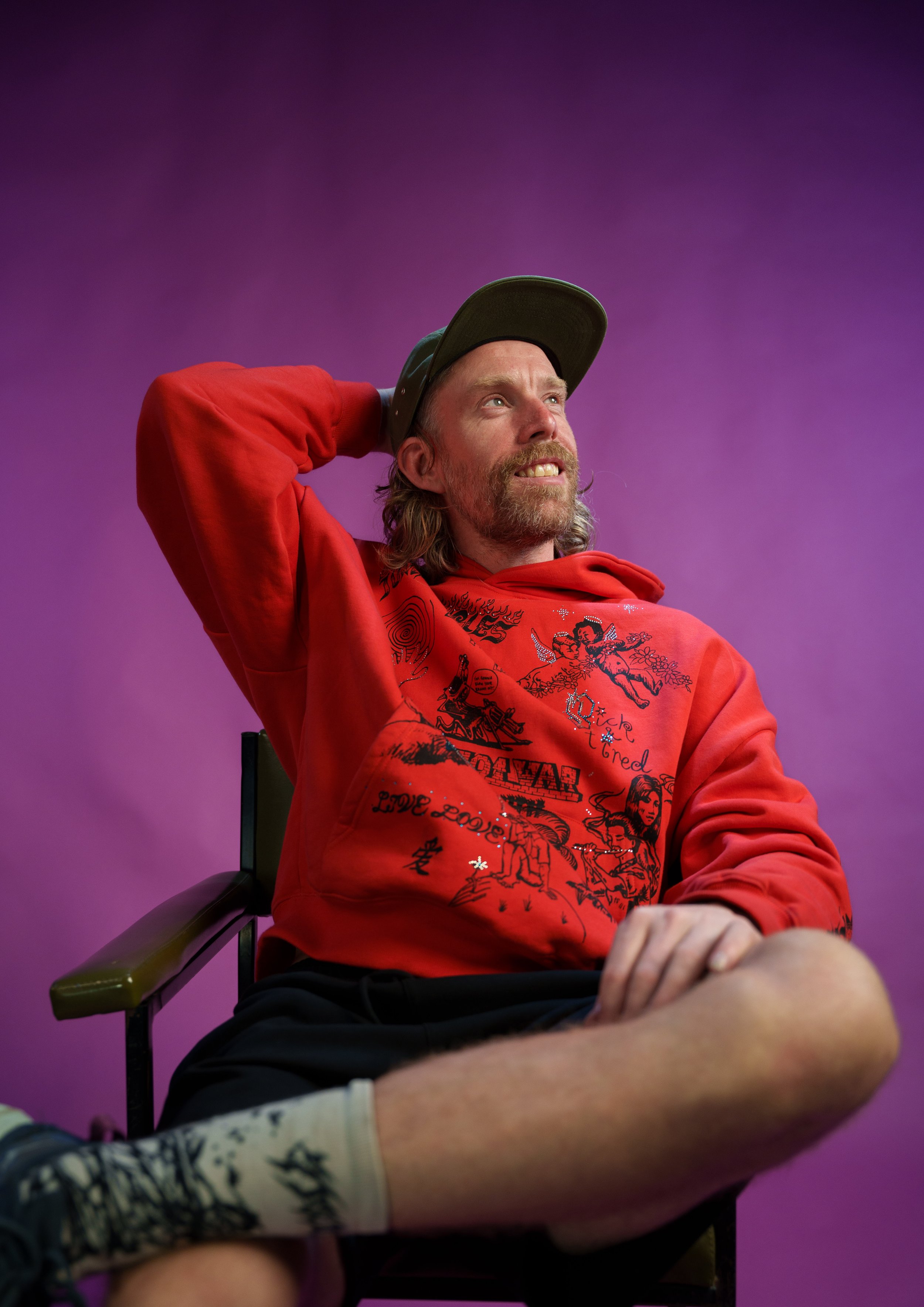 A man with long hair and a beard wearing a black cap and red hoodie, sitting in a chair with a purple background, smiling and looking up.