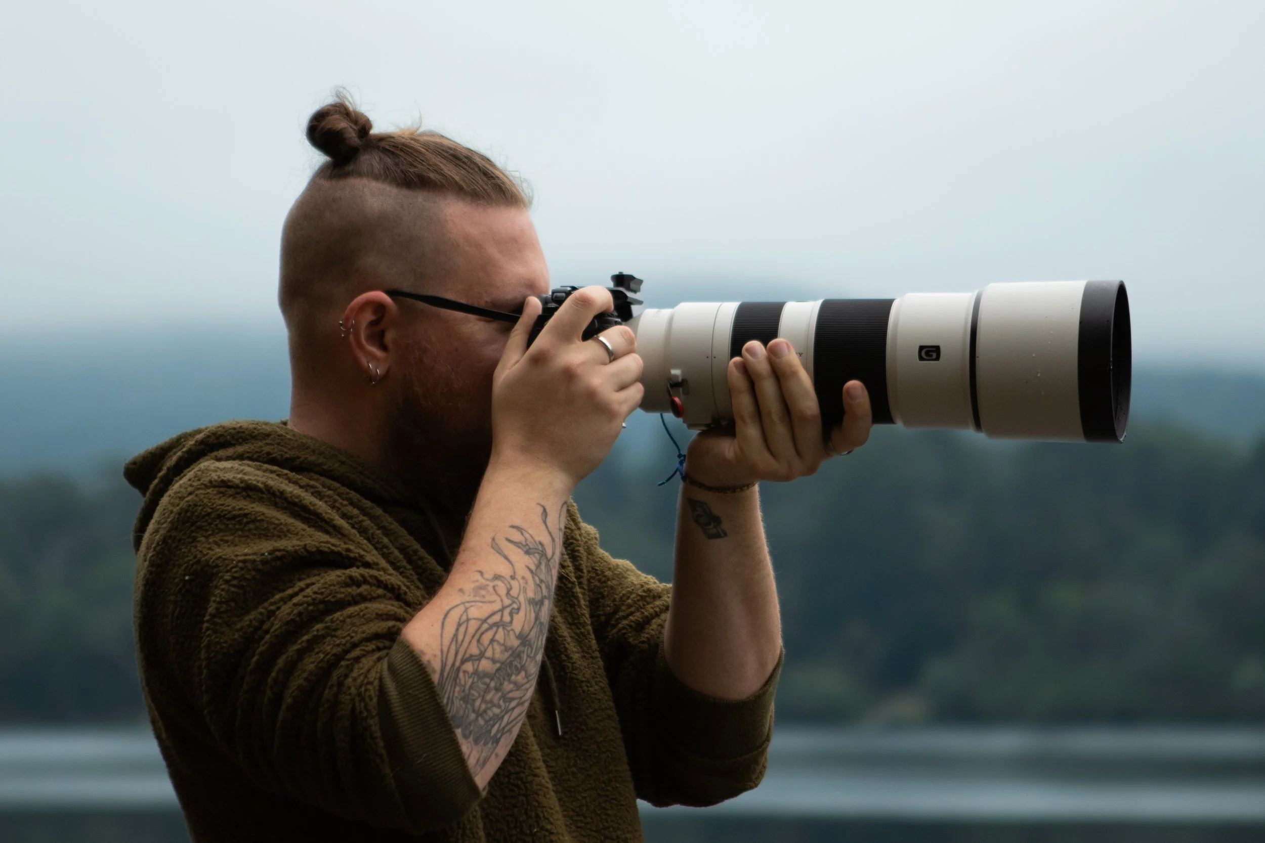 Man with tattoos and glasses taking a photo with a long telephoto lens camera outdoors.