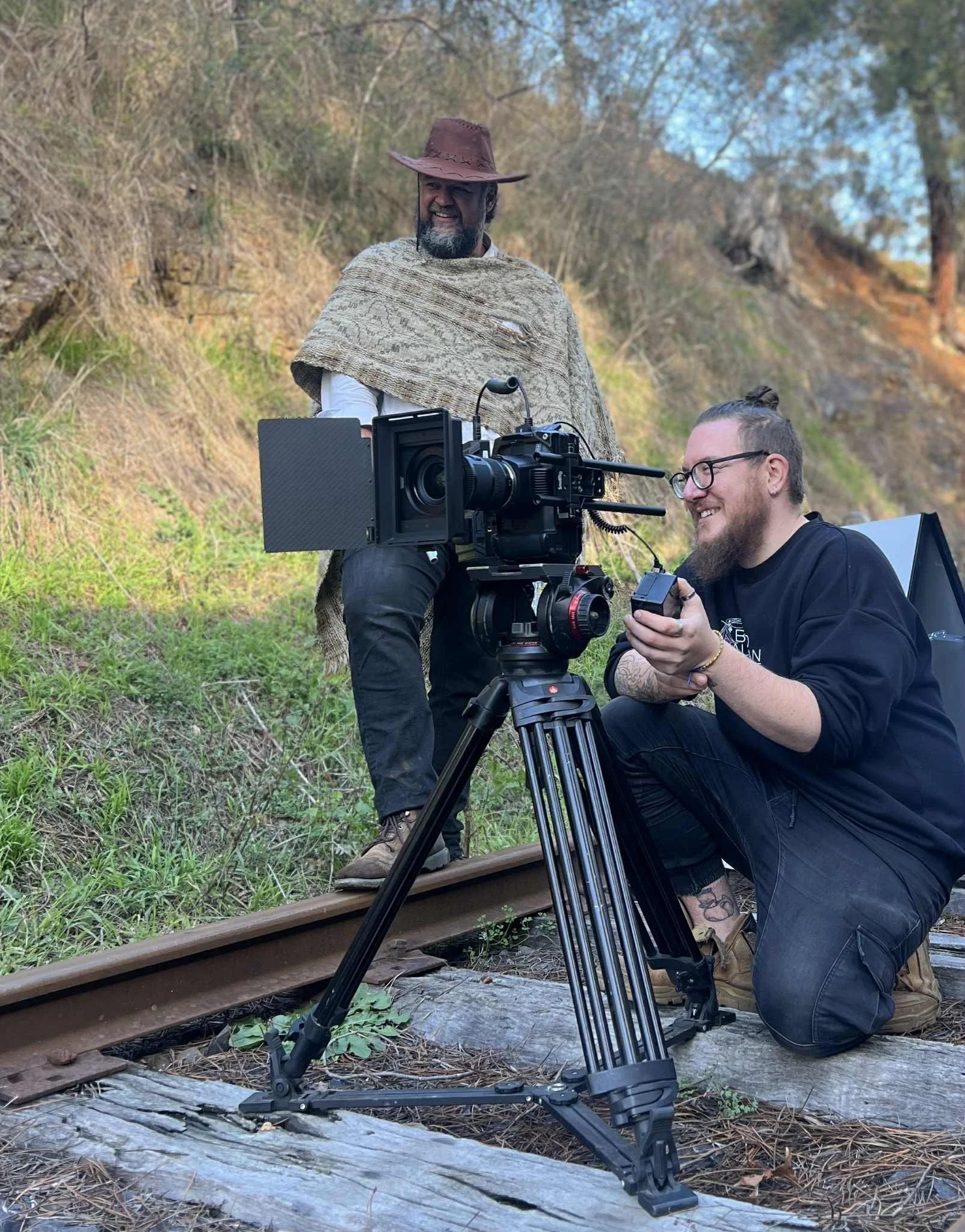 Two men on a railroad track with a professional film camera, one standing in cowboy costume with a poncho and wide-brimmed hat, the other kneeling with glasses and a tattoo, adjusting camera settings.