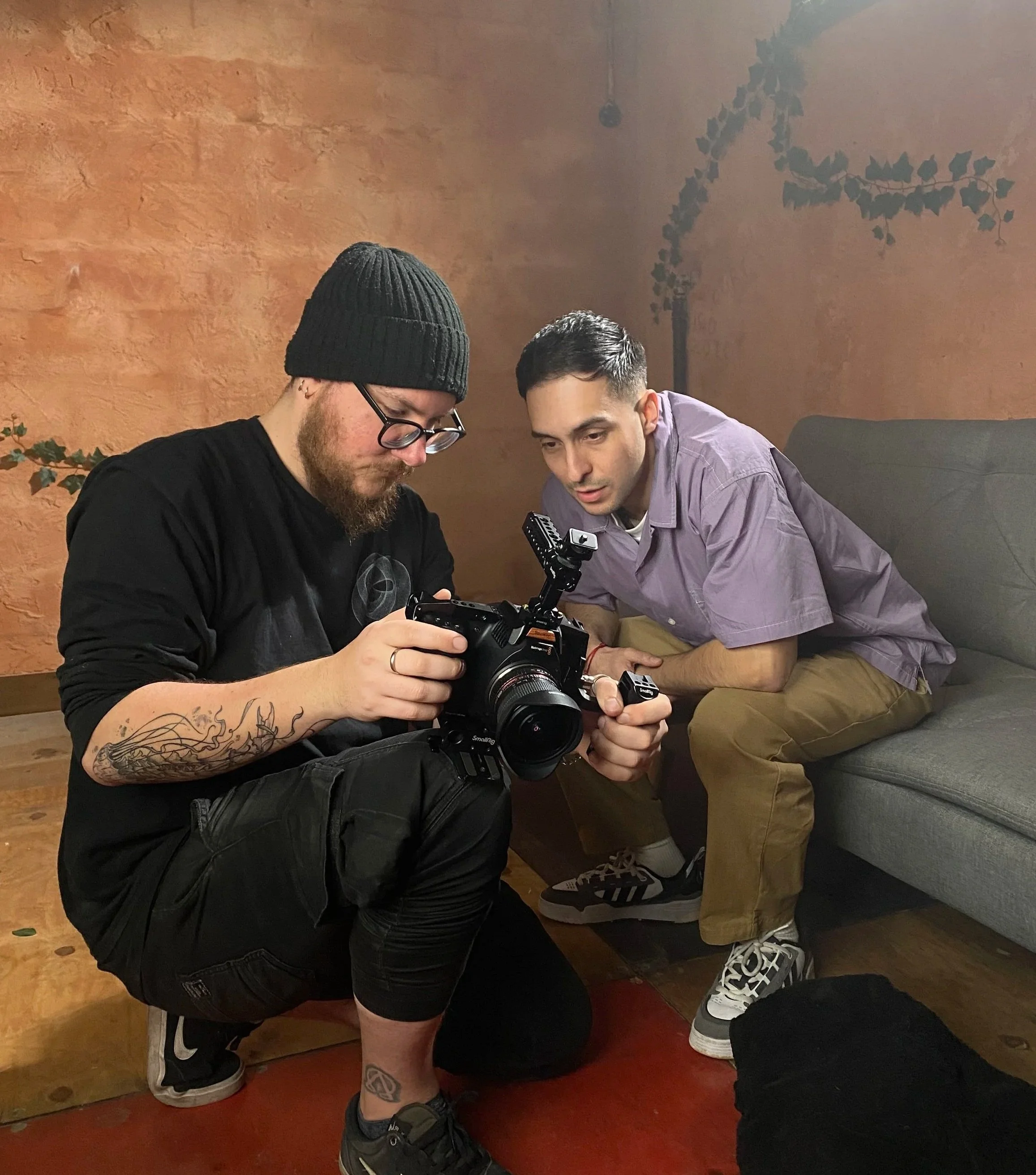 Two men examining a professional camera together in a cozy room with an orange wall and some decorative vines.