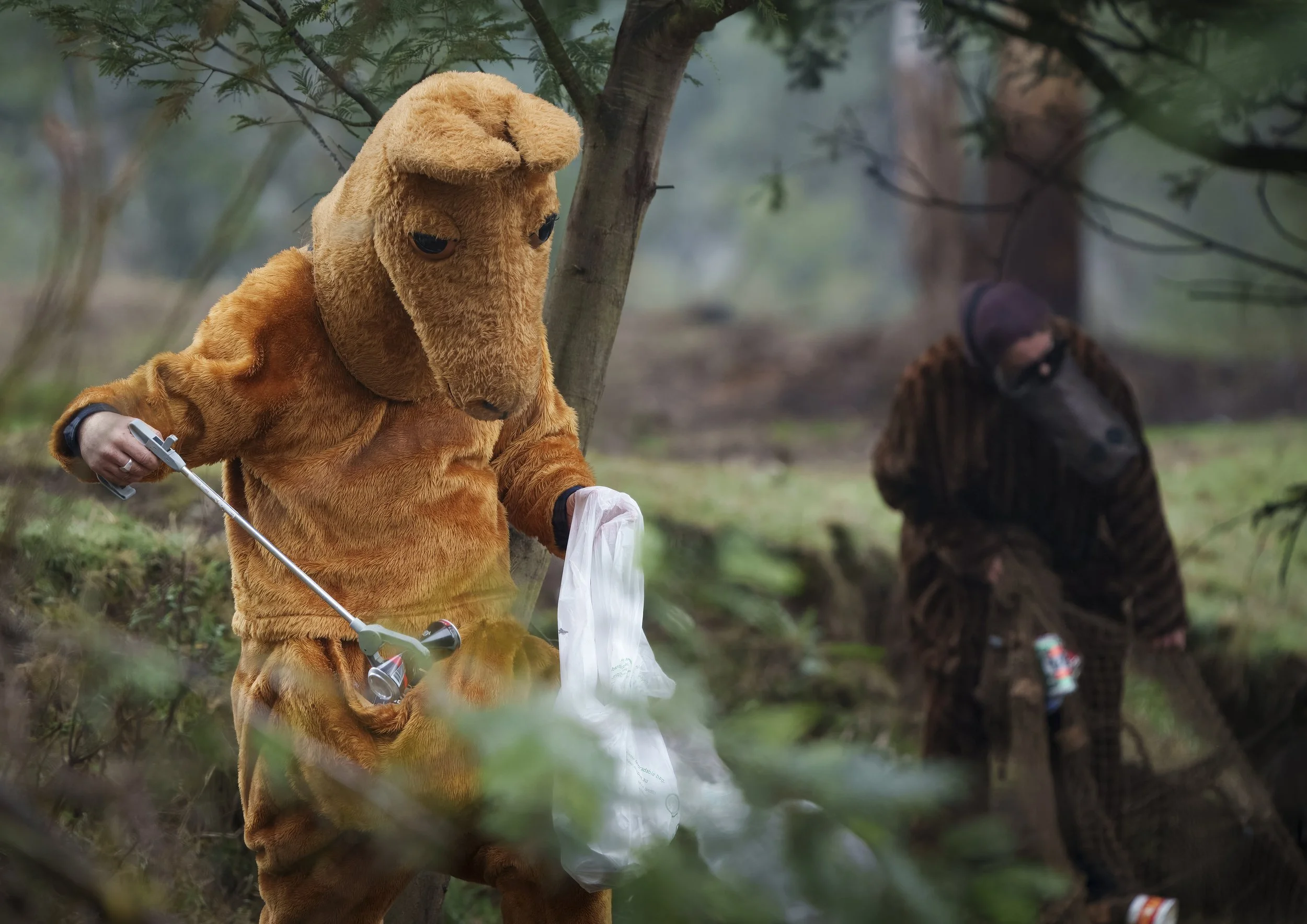 A person in a kangaroo costume holding a plastic bag, collecting rubbish, outdoors among trees.
