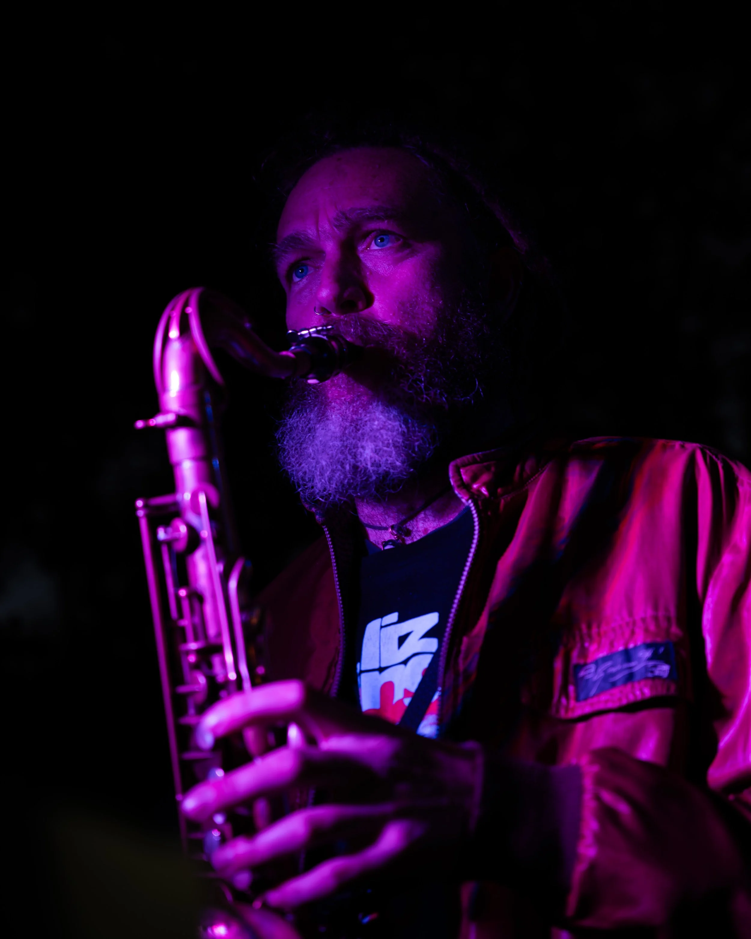A person with blue eyes and a beard playing a saxophone in a dark setting illuminated by purple lighting.
