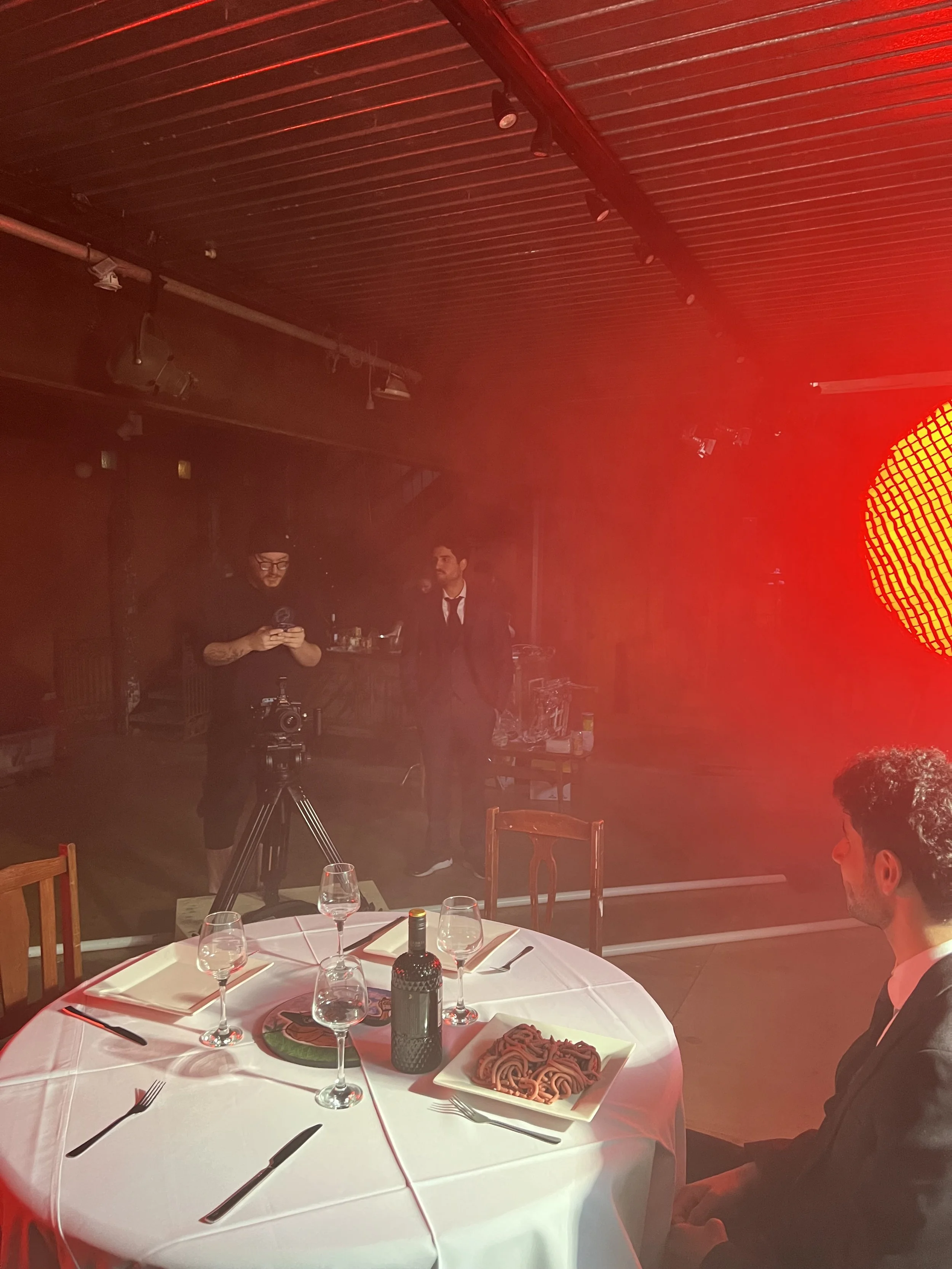 A man in a suit sitting at a round dining table with wine glasses, a bottle of red wine, and plates of meat, with two men in suits, a camera, and a large red light in the background.