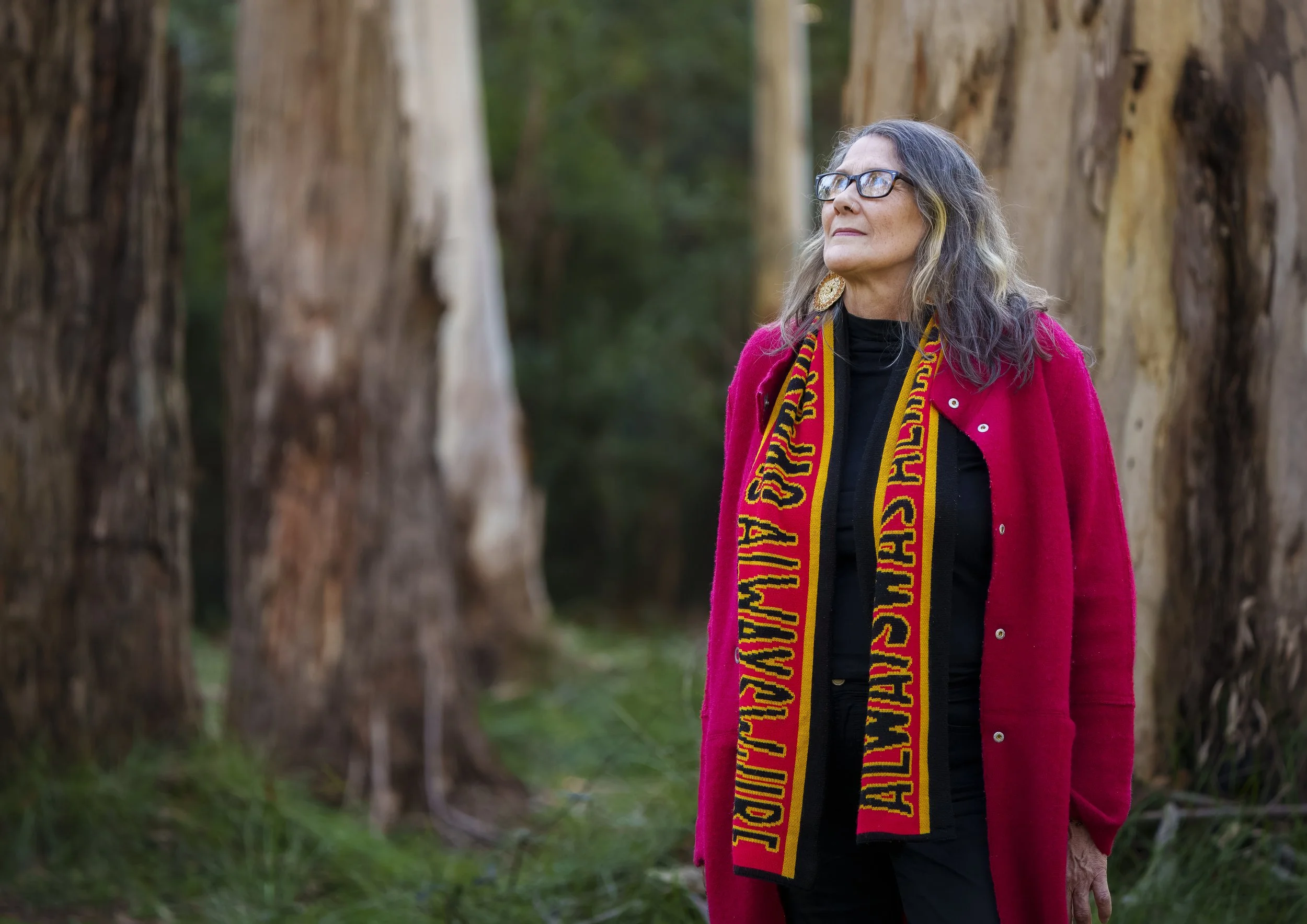 An indigenous elder woman with grey hair and glasses, wearing a red jacket and a yellow and red scarf that says 'always was, always will be' stands outdoors among tall trees, looking off into the distance.